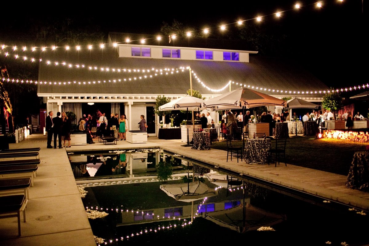 Crisscrossed café string lights glow over a reflecting pool and mingling guests at an evening wedding reception lit by Impact at Durham Ranch.
