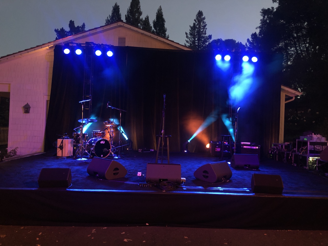An outdoor backyard performance stage set with drums, PA speakers and blue wash lighting against black drape at dusk, staged by Impact.