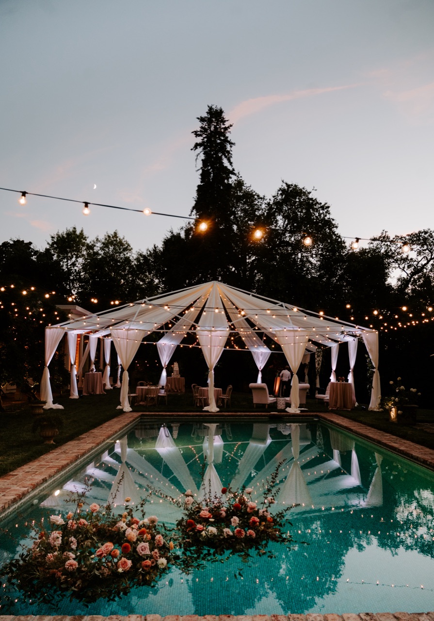 Impact lights a clear-top pole tent and surrounding trees with warm bistro string lights over a reflecting pool at a dusk garden wedding.