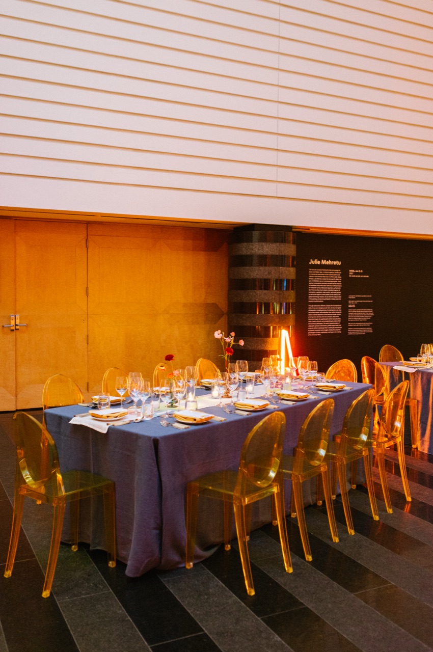 An intimate museum dinner at SFMOMA with amber ghost chairs and a lavender table beside a gallery text wall, styled by Impact.