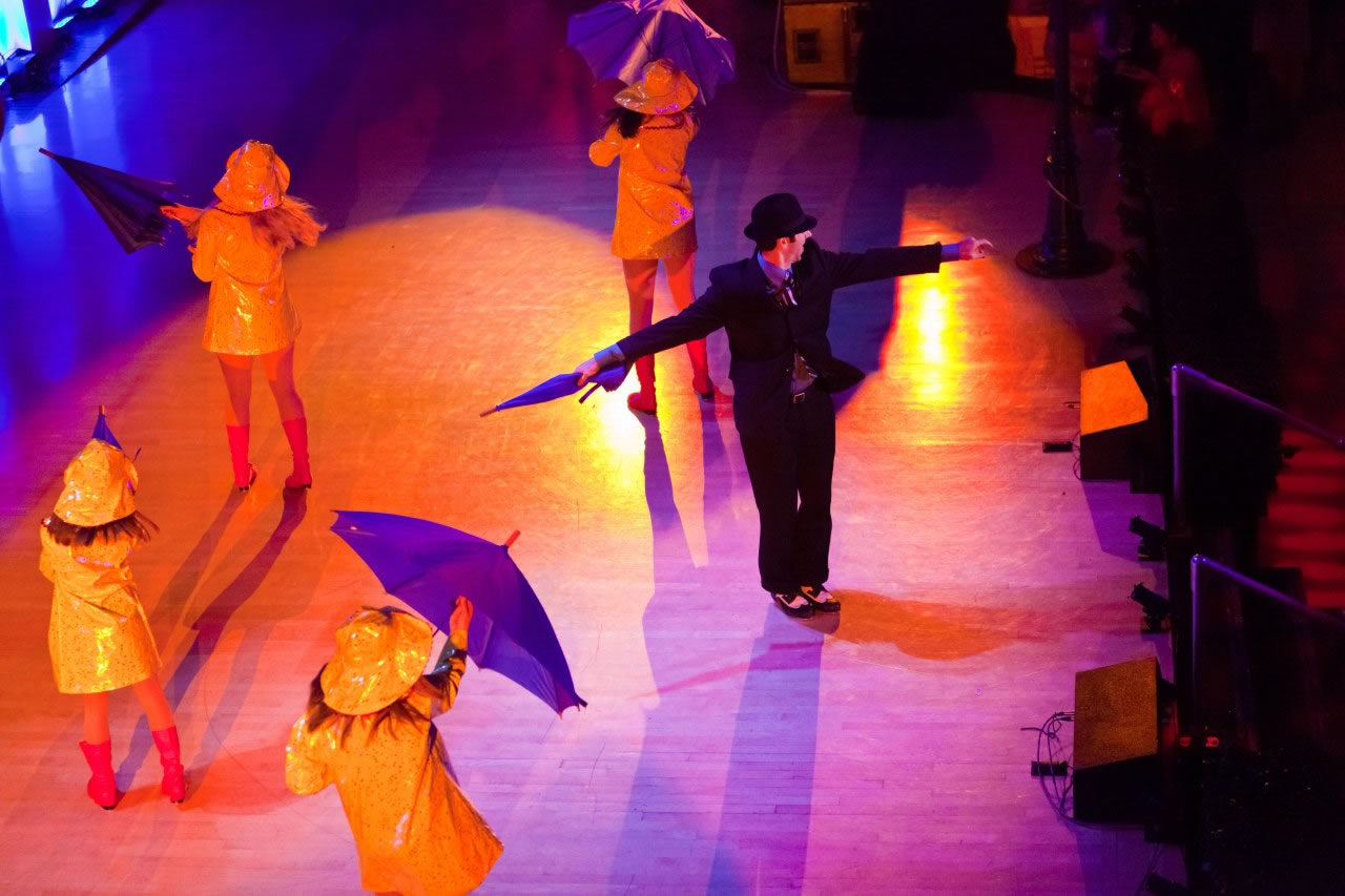 A themed stage performance of dancers in yellow raincoats with umbrellas under amber and purple floor light, produced by Impact.