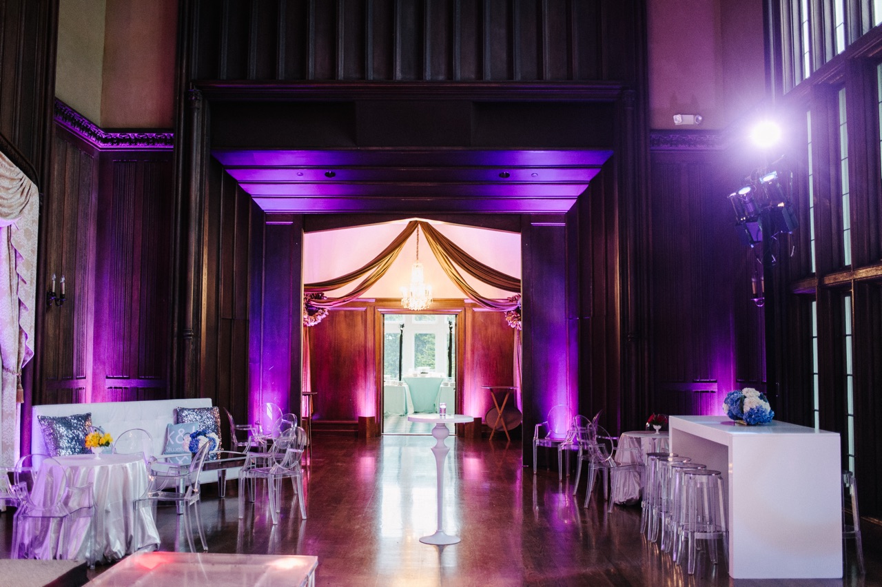 A dark wood-paneled hall with purple uplighting, a draped chandelier-lit doorway and ghost-chair lounge furniture styled by Impact for a bat mitzvah.