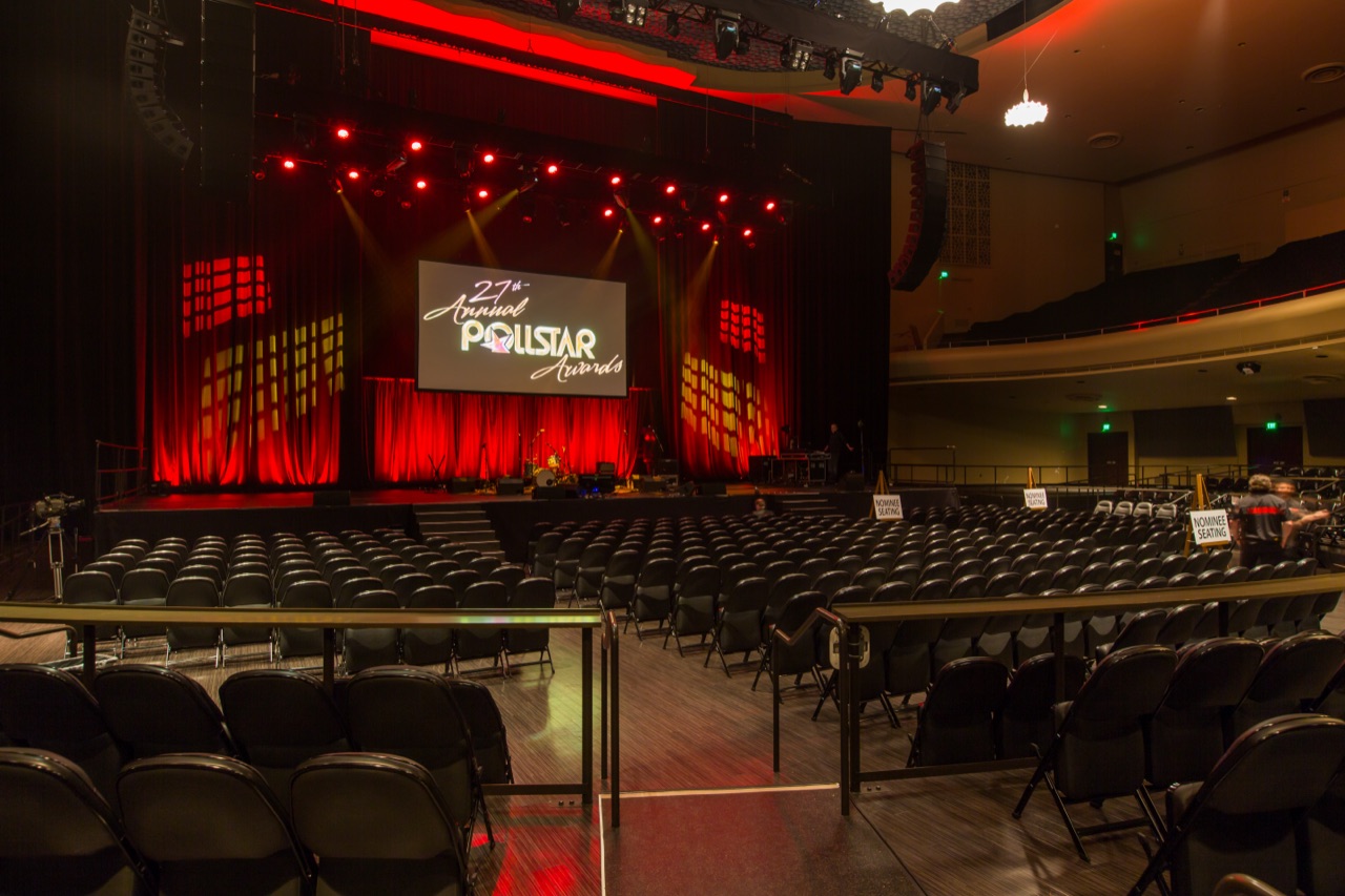 A red-washed concert stage with beam lighting and the Pollstar Awards screen viewed from the side of the empty house, lit by Impact.