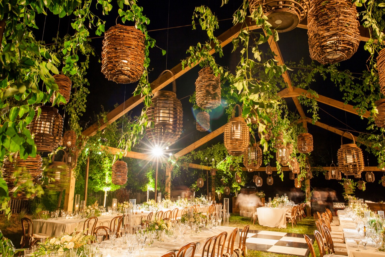 An outdoor wedding dinner glows under a pergola of hanging woven lanterns, trailing greenery and warm market lighting by Impact at Gardner Ranch.