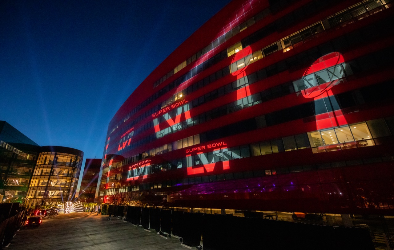 Impact projected Super Bowl LVI graphics across a curved building facade in scarlet and gold at dusk.