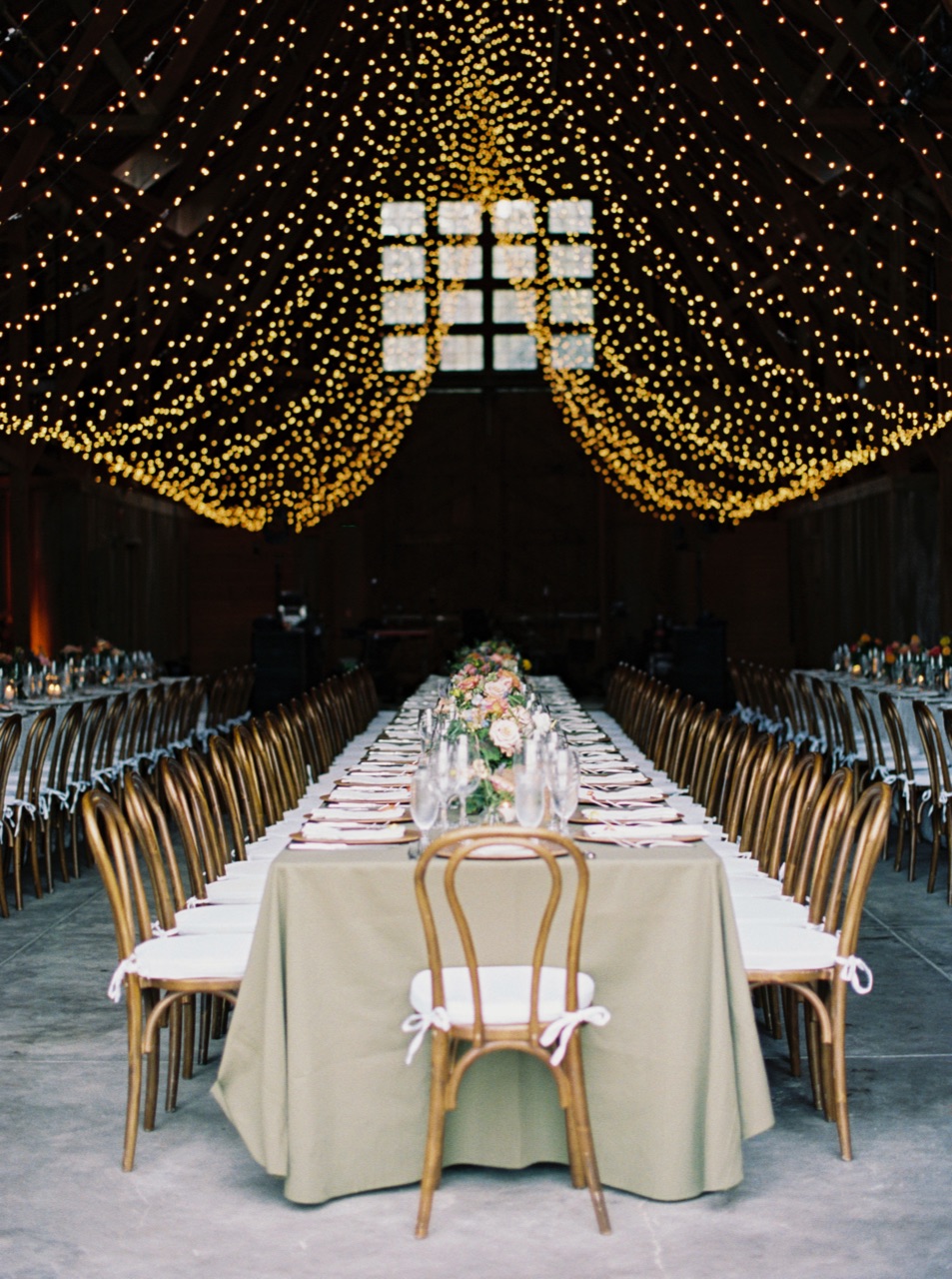 Impact suspends a dense canopy of warm fairy lights across a barn ceiling above a long banquet table with cross-back chairs at a Santa Lucia Preserve wedding.