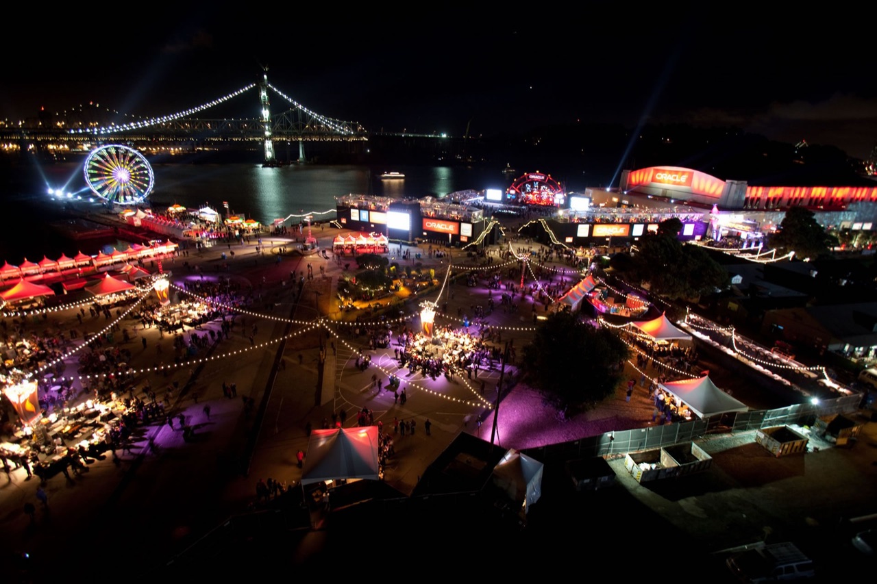 An aerial night view of Impact's waterfront festival lighting with festoon strings, tents, and a stage beneath the Bay Bridge.