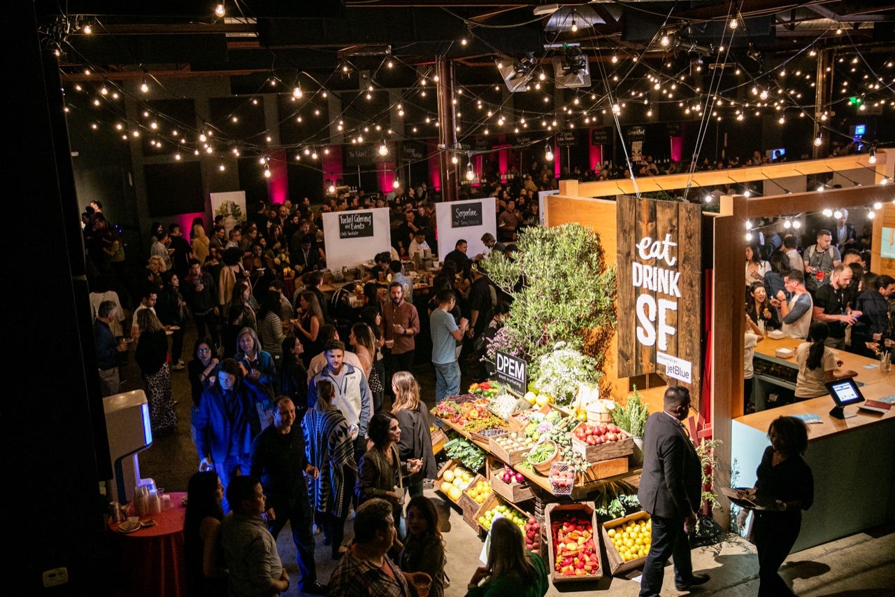 Edison-bulb festoon strings and red uplighting hang above the crowd and a produce display at Impact's Eat Drink SF event.
