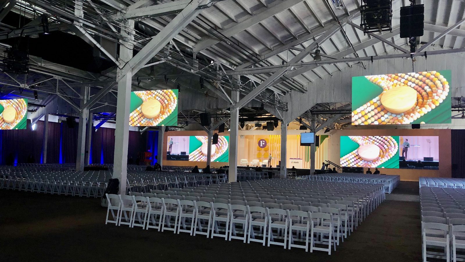 A cavernous hangar-style event space fitted by Impact with rows of synchronized LED video walls flanking a keynote stage and chairs set for a corporate general session.