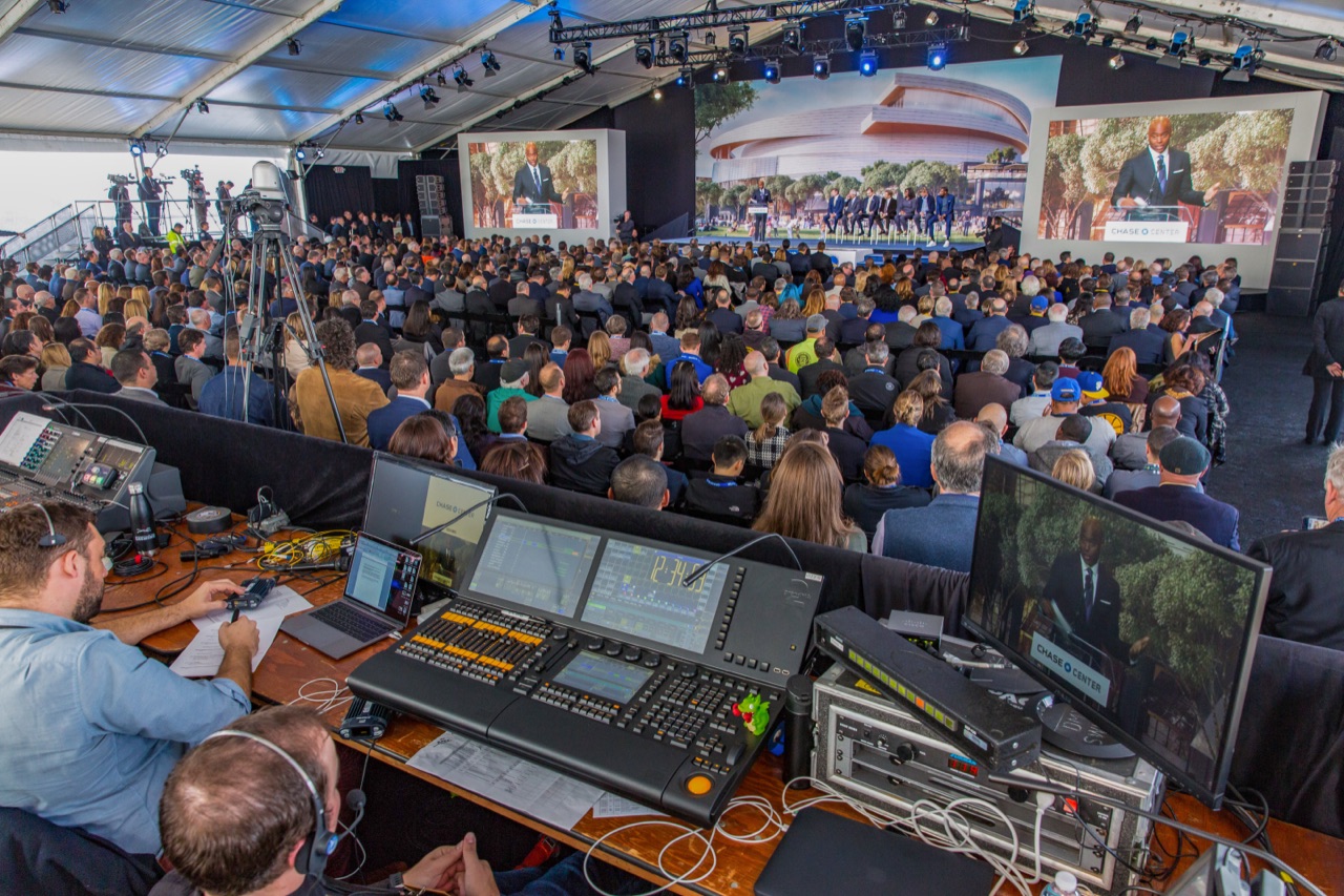 A lighting console anchors the foreground as a crowd watches a keynote on large LED screens at a corporate groundbreaking produced by Impact.