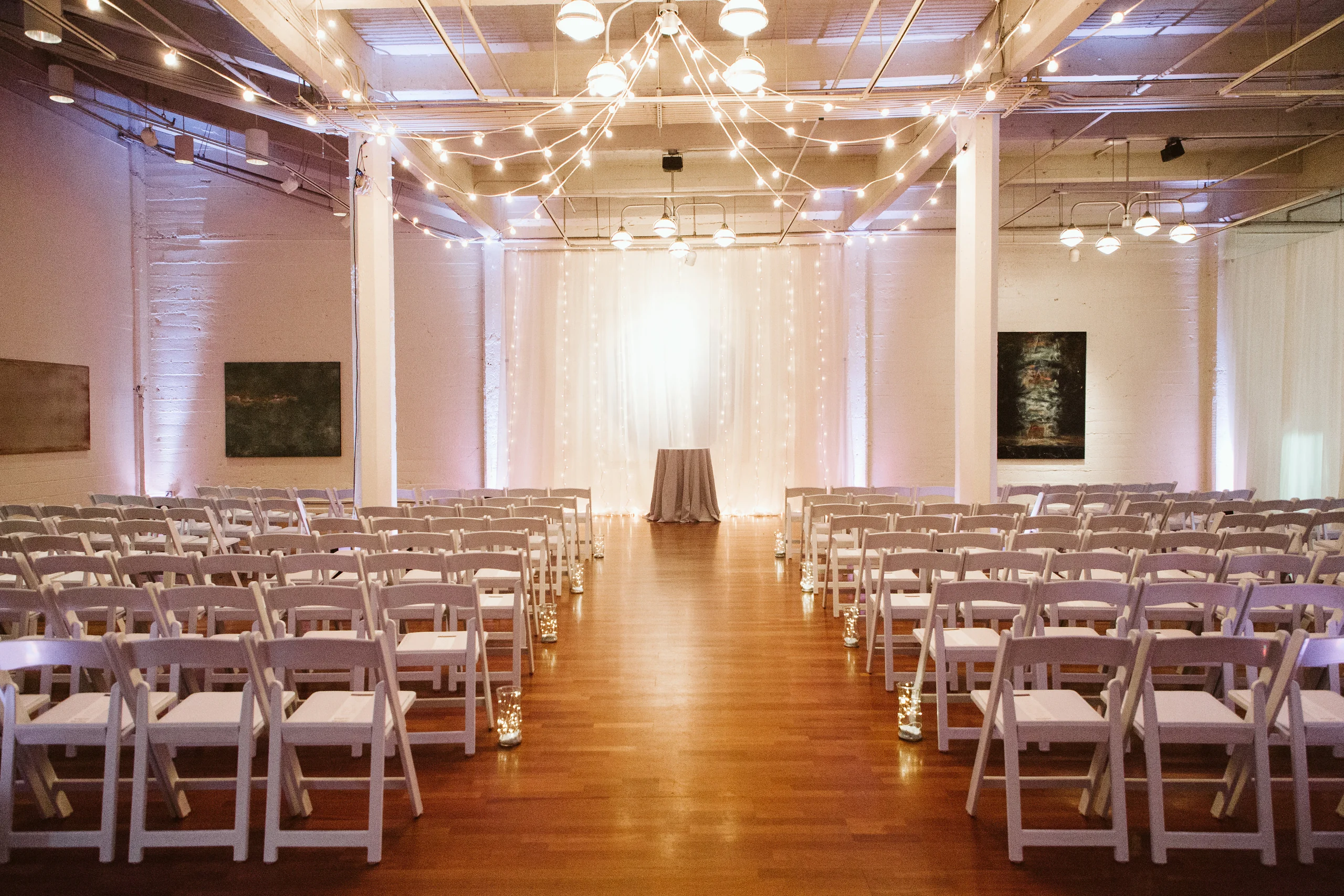 Impact lines a loft gallery ceremony with crisscrossed string lights, soft pink uplighting on the columns, and a fairy-light backdrop down the aisle at Terra Gallery.
