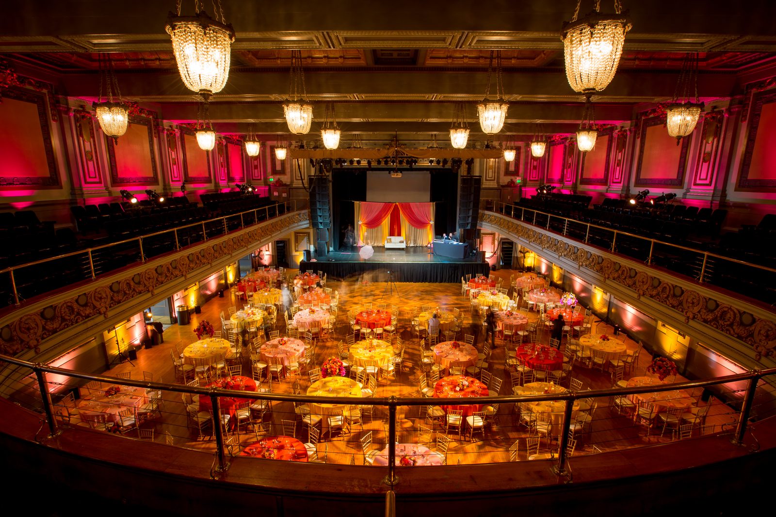 Impact lights the Regency Ballroom from the balcony with amber and red uplighting, crystal chandeliers, and a floral stage mandap above colorful banquet tables at a South Asian wedding.