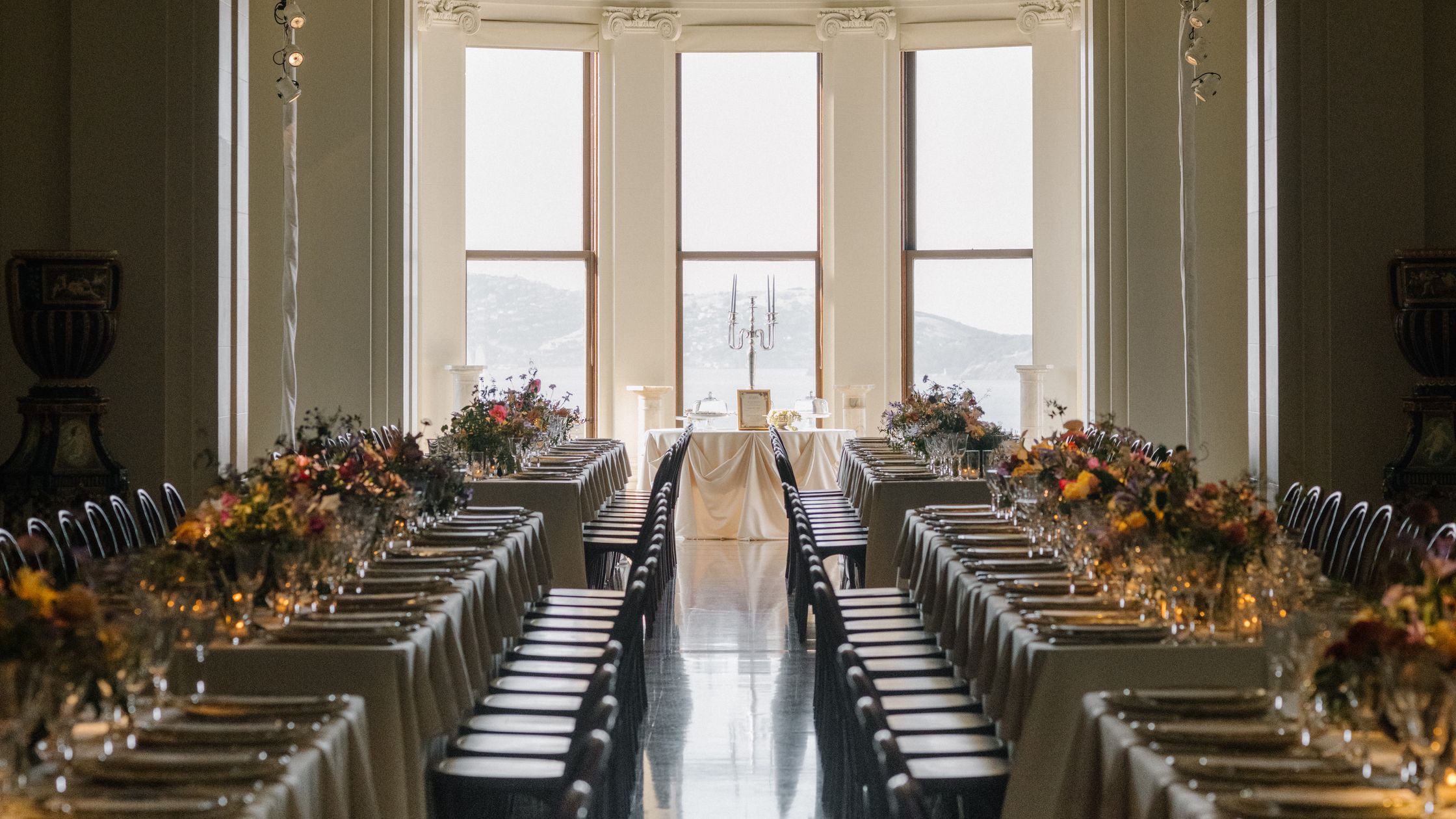 Long reception tables set with autumn florals and candelabras in a grand hall with tall arched windows