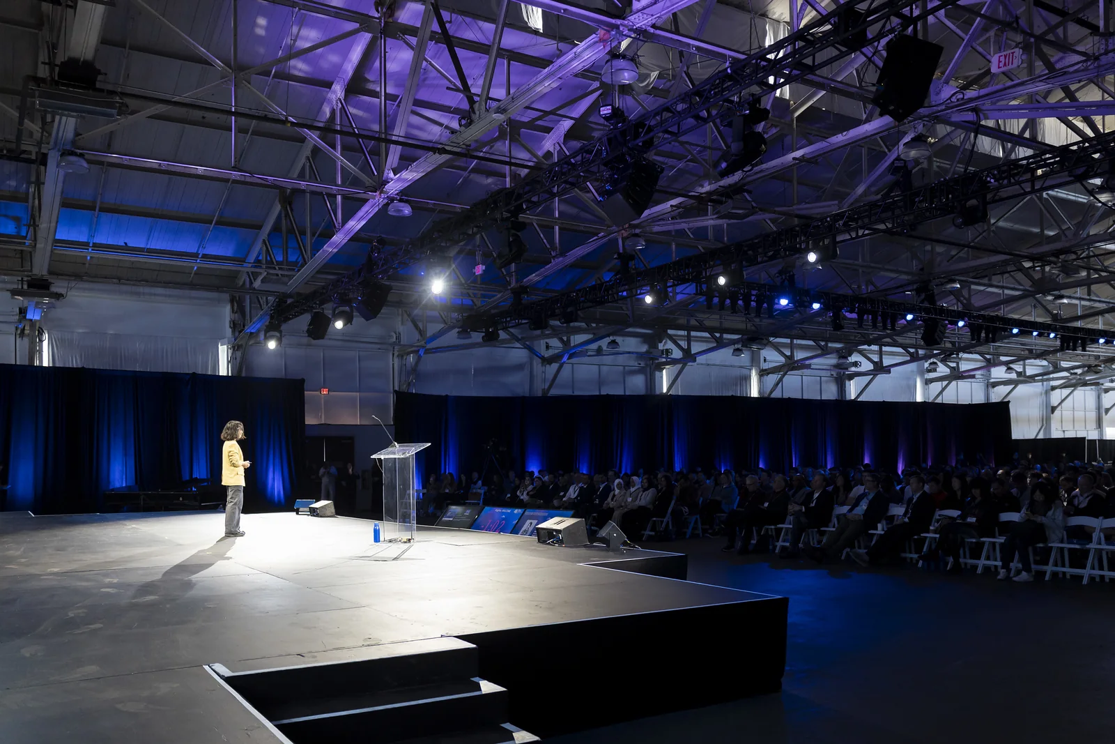 A presenter on a keynote stage under a truss lighting rig, with blue-washed drape behind the audience