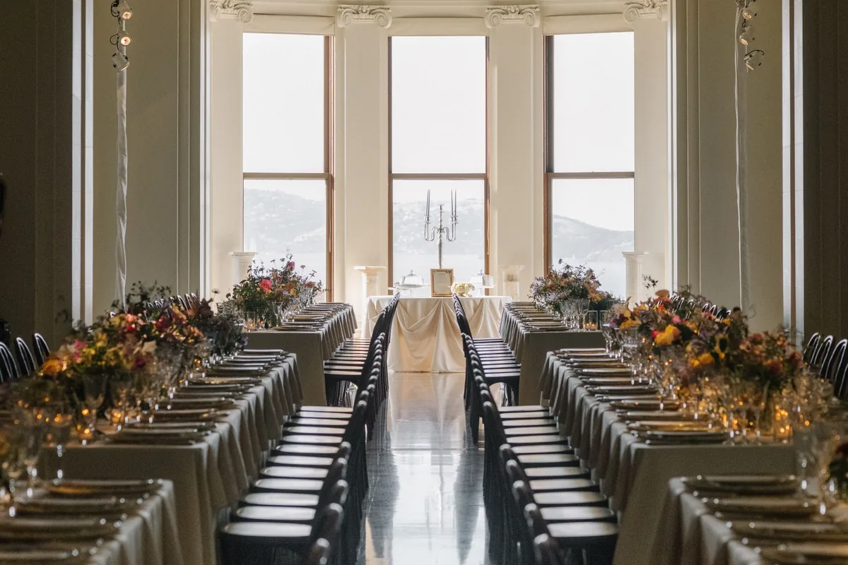 Long reception tables set with autumn florals and candelabras in a grand hall with tall arched windows