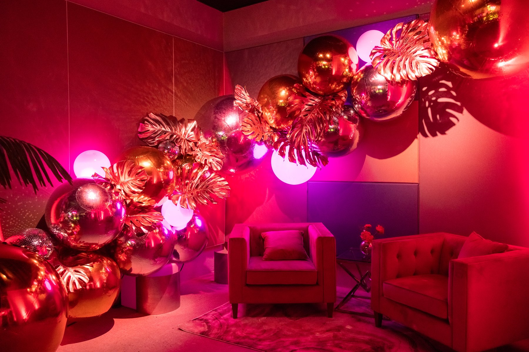 Pink velvet armchairs beneath an installation of mirrored rose-gold orbs and gilded palm leaves, washed in magenta light.