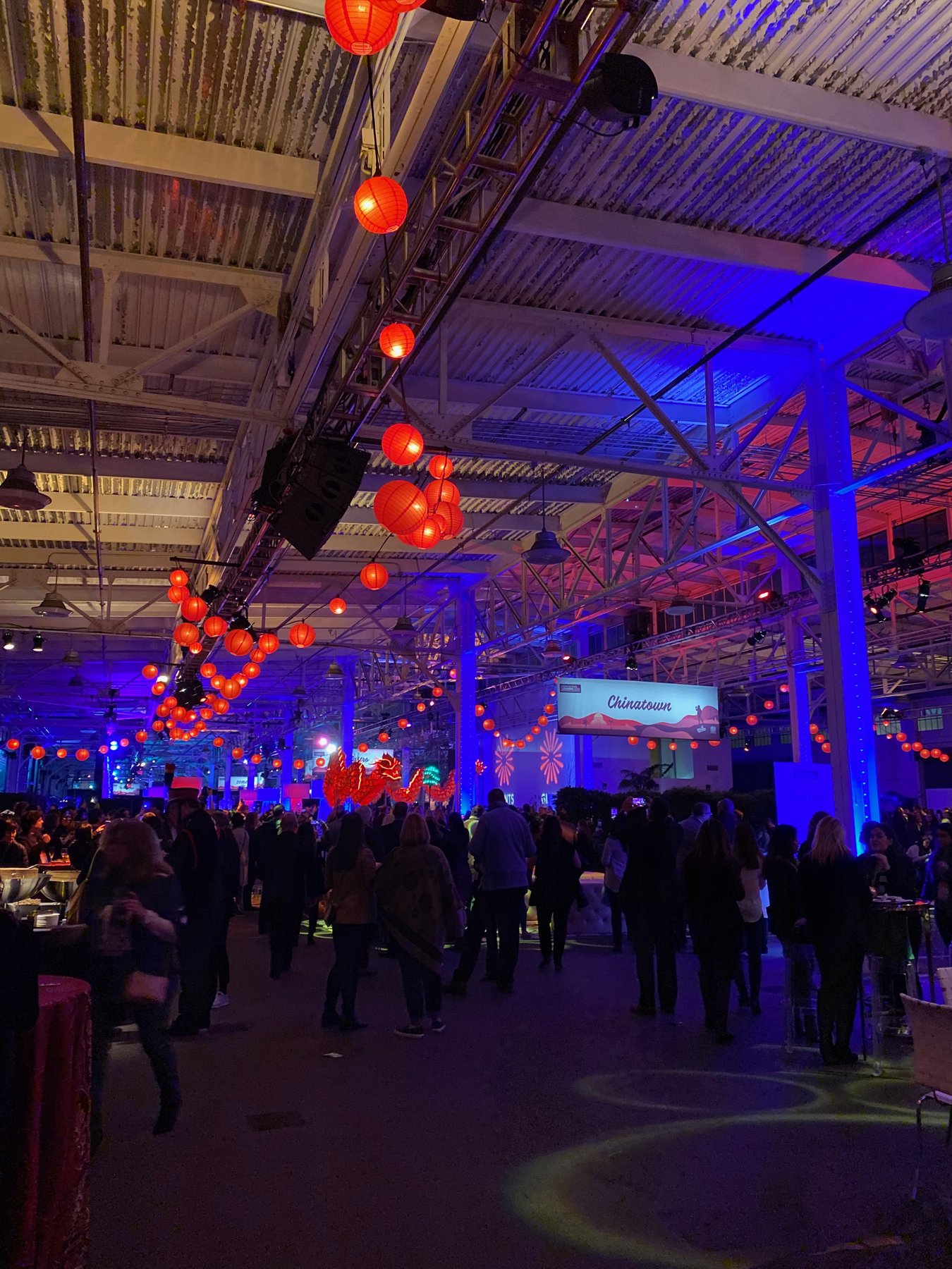 Strings of red paper lanterns hang from truss above a crowded reception in an industrial hall washed in blue light.