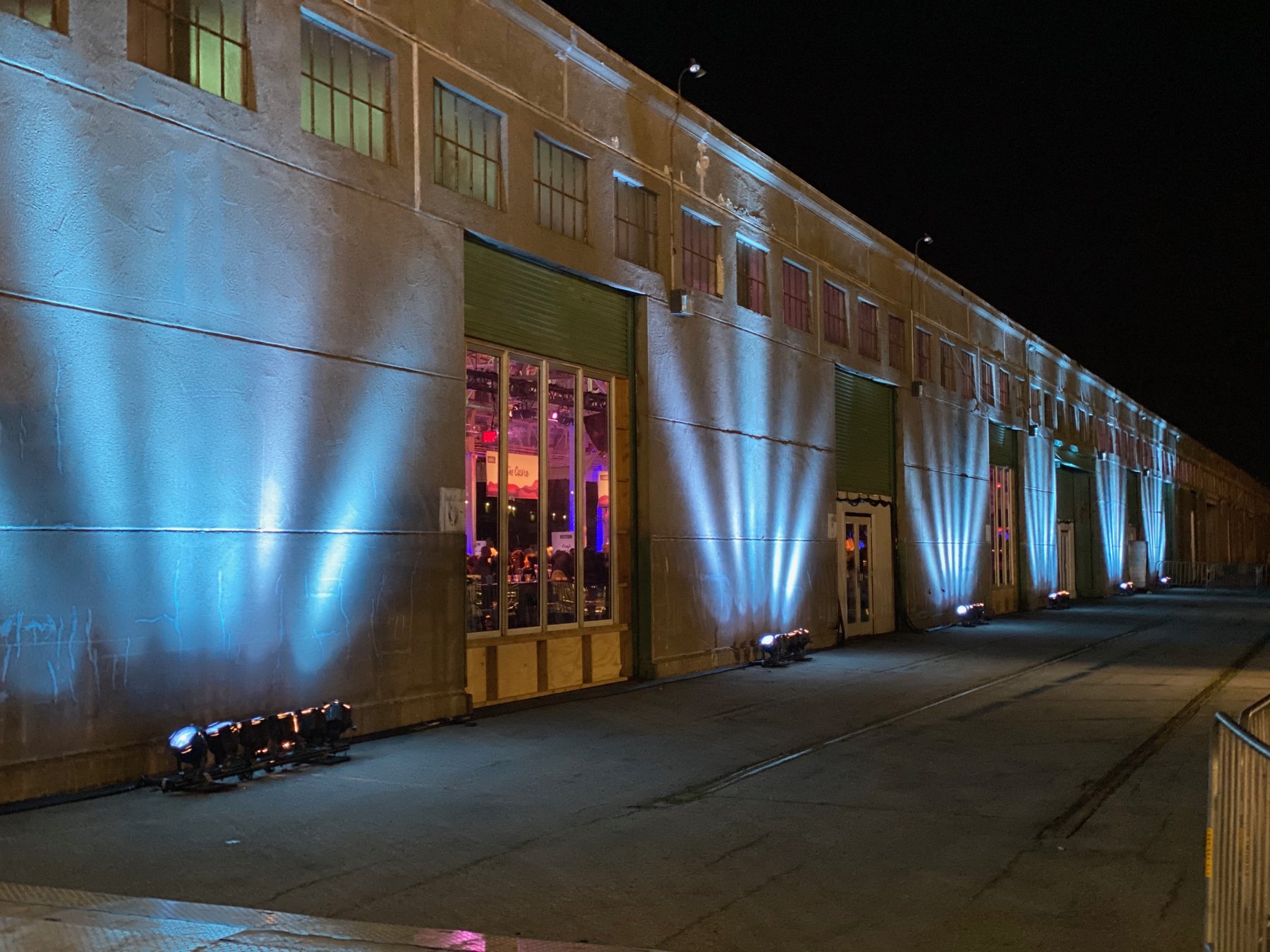 Fans of pale blue uplight climb the exterior wall of a long warehouse at night, with the event visible through an open door.