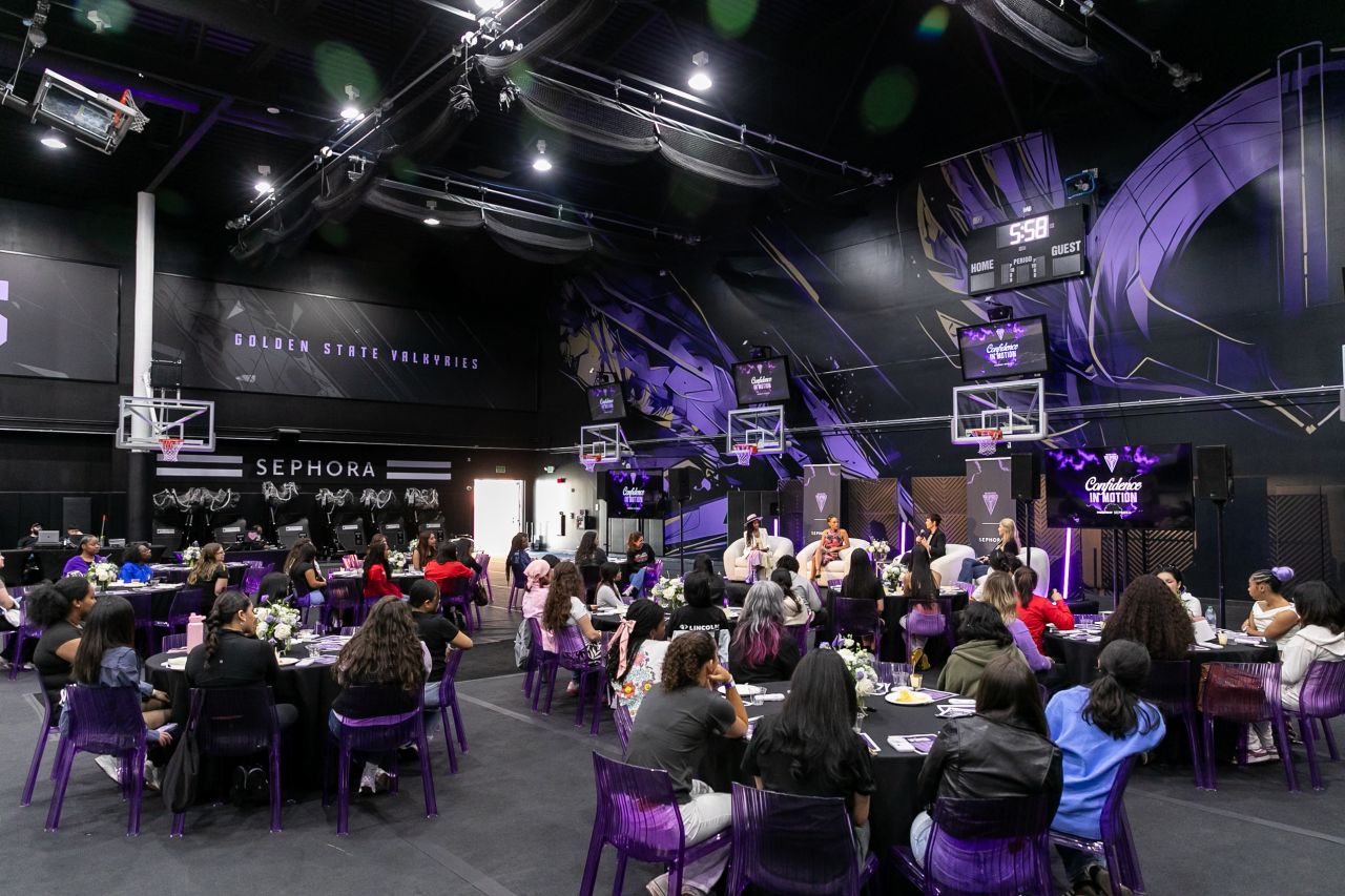 Guests at round tables with violet chairs watch a panel inside a basketball practice facility, with hoops overhead and violet light on the walls.