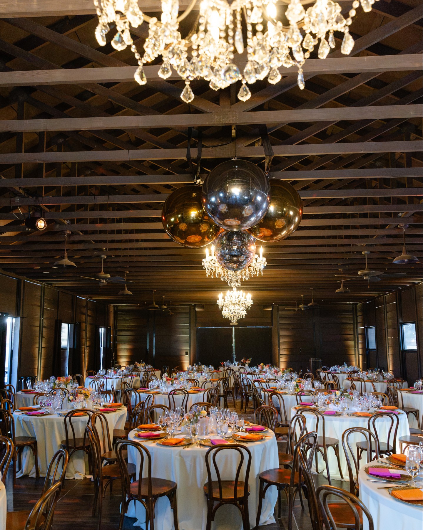 A crystal chandelier and a cluster of mirror balls hang from the rafters of a timber barn above round dinner tables with bentwood chairs.
