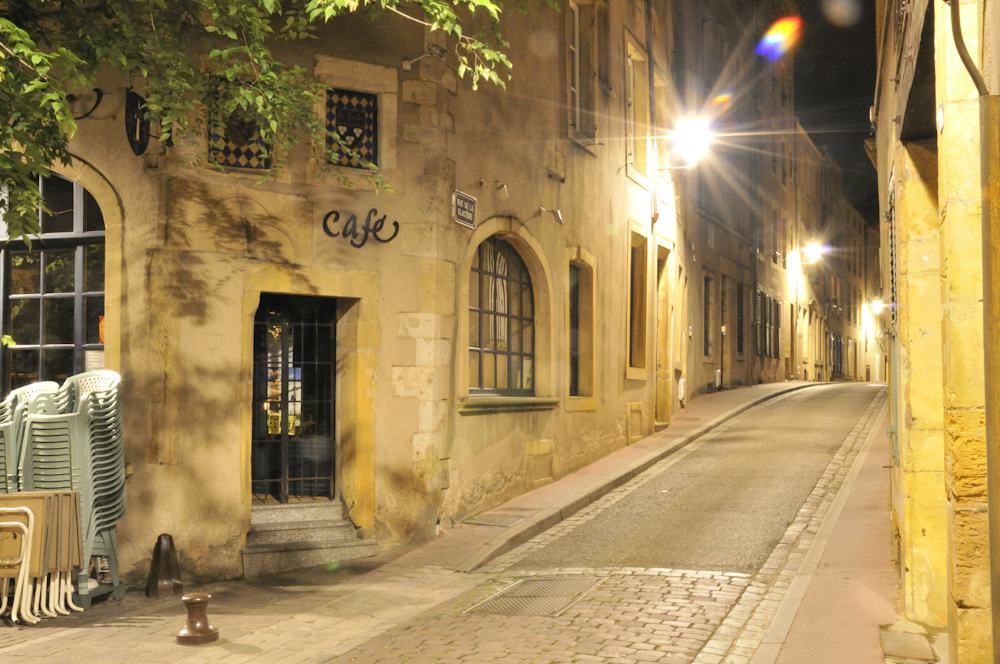 Une ruelle pavée de centre ancien la nuit, éclairée par des réverbères, bordée de maisons en pierre.