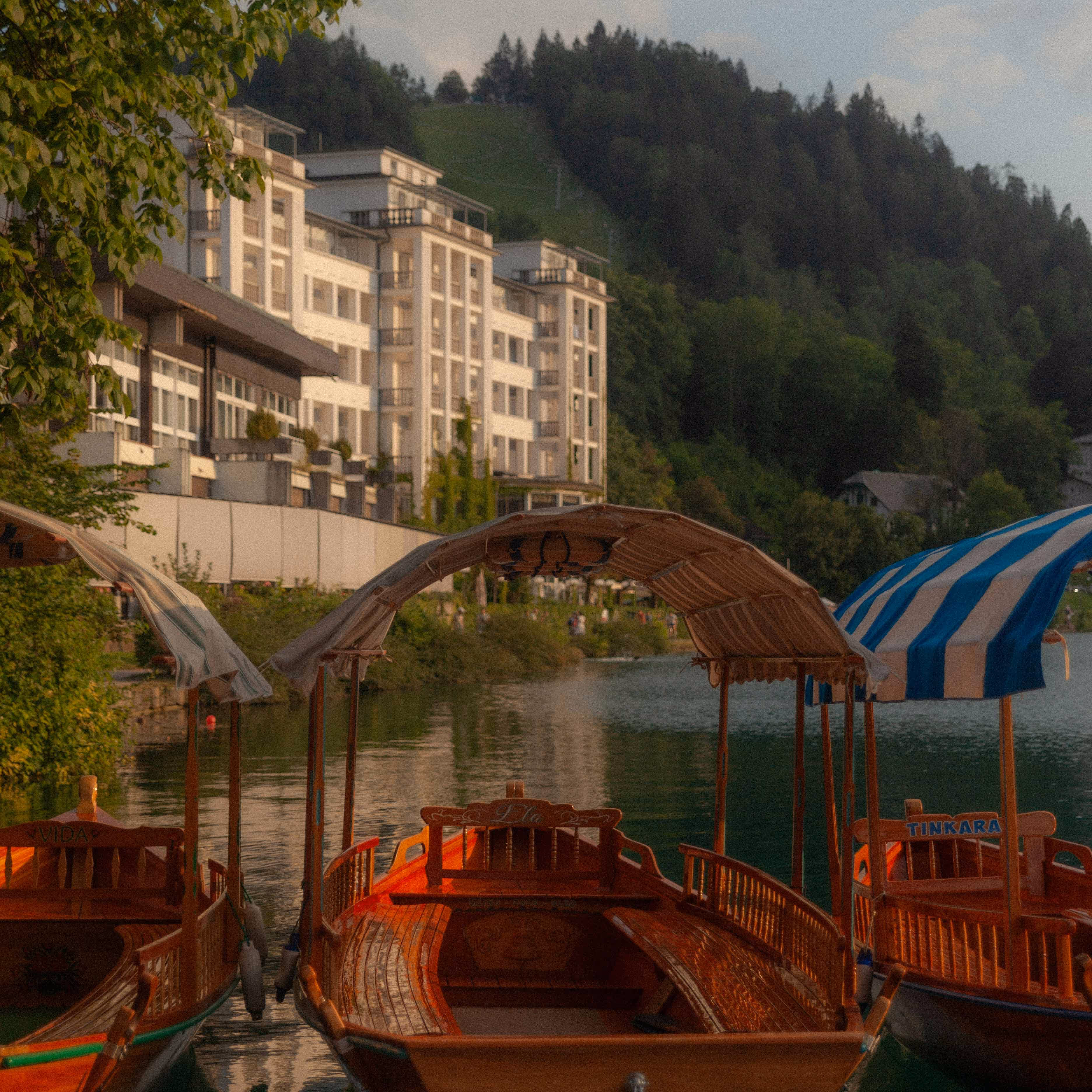 Three wooden boats with canopies docked at a calm lake with a luxury hotel and forested hills in the background.