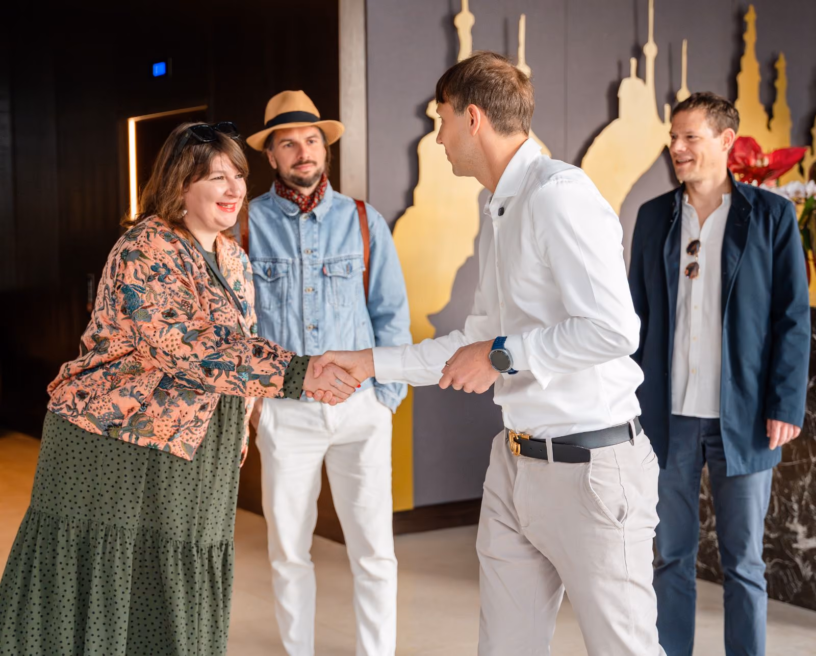 A woman in a floral patterned jacket shakes hands with a man wearing a white shirt and beige pants, while two other men look on smiling, one dressed casually with a hat and denim jacket and the other in a dark blazer and white shirt.
Meet and greet in the hotel in Ljubljana