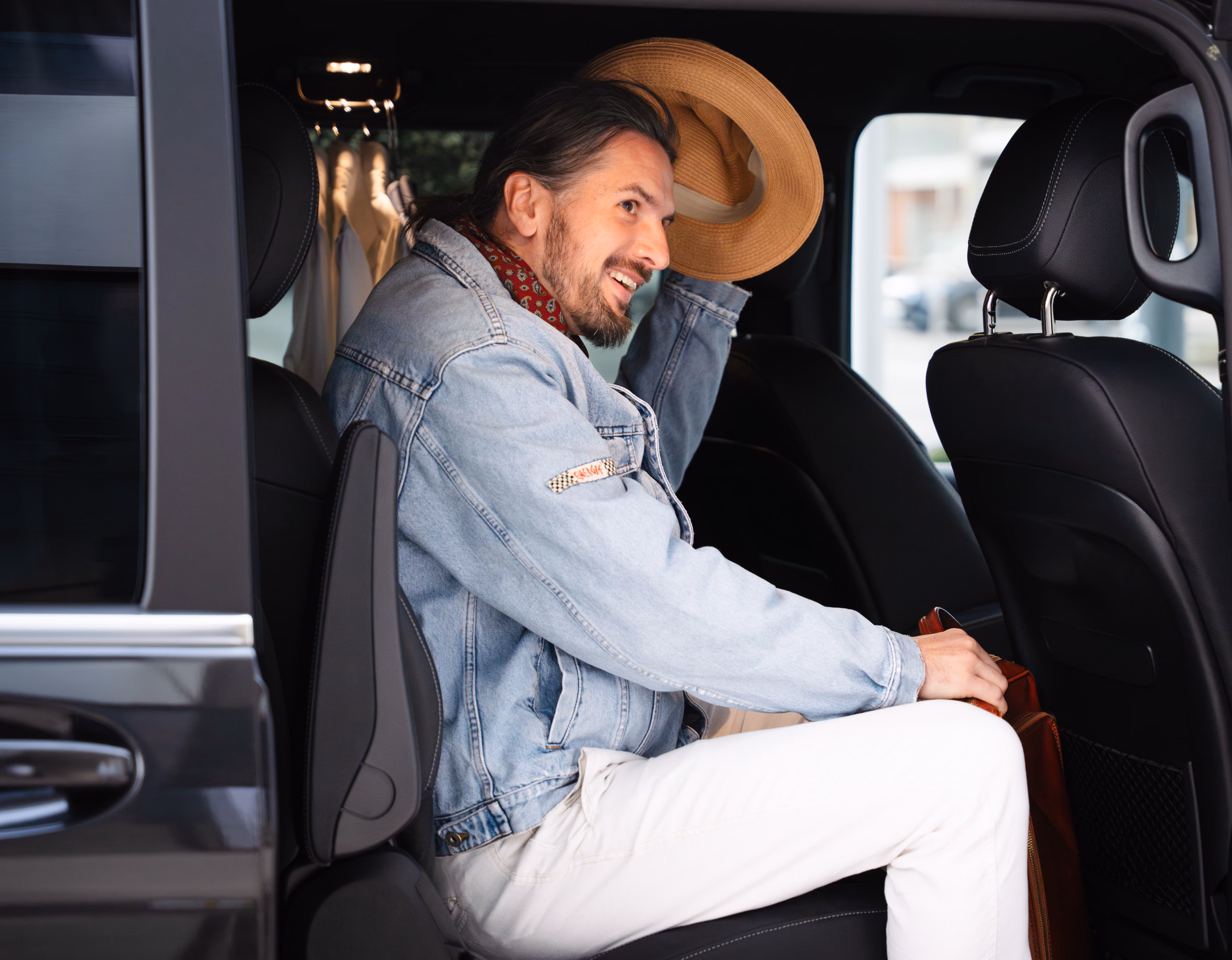 Man sitting inside a black vehicle smiling and holding a tan hat with one hand, wearing a light denim jacket, a red neckerchief, and white pants.
Man takeing goodbay