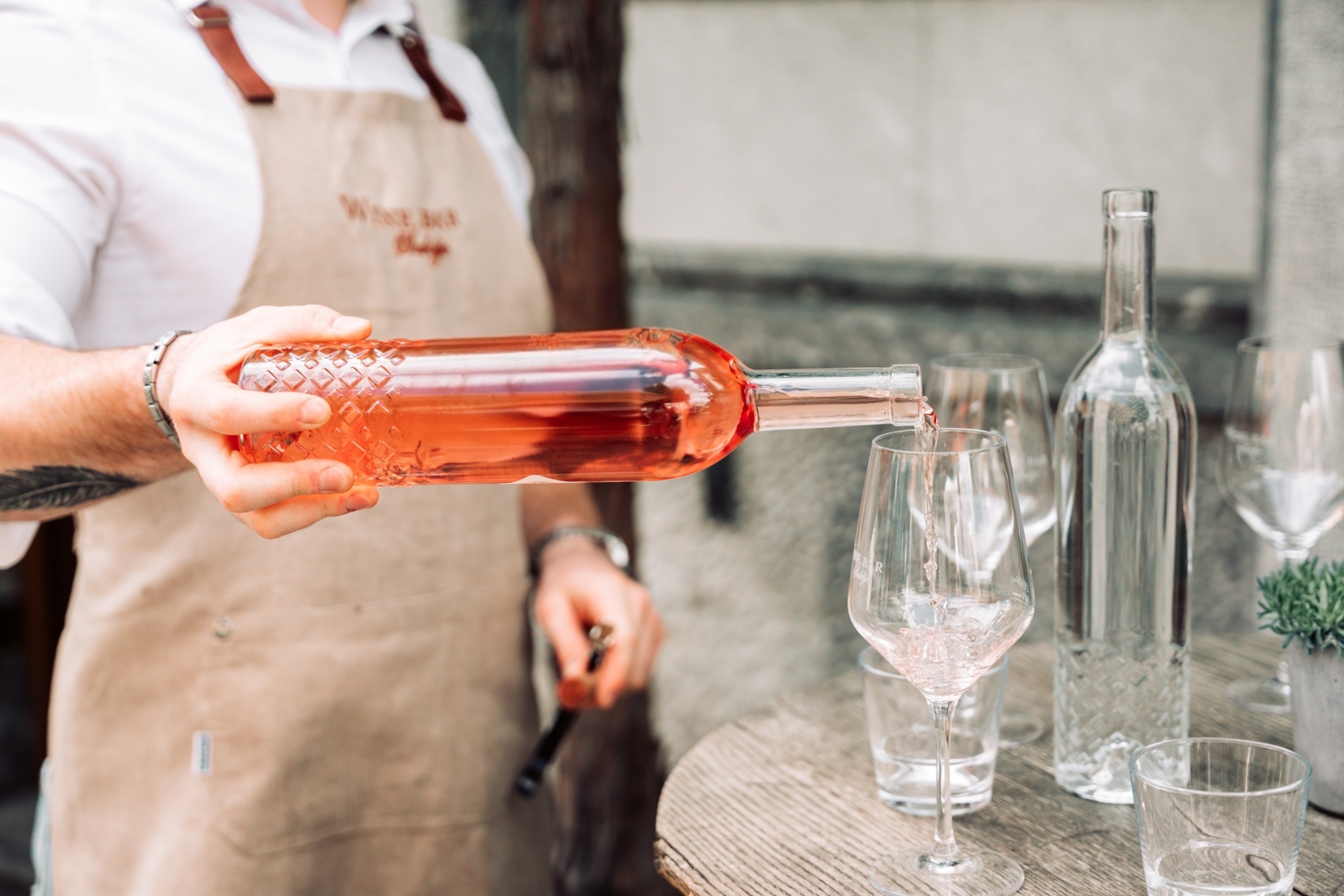 A person in a beige apron pouring rosé wine from a textured glass bottle into a clear wine glass set on a wooden table, alongside a bottle of water and additional glasses.