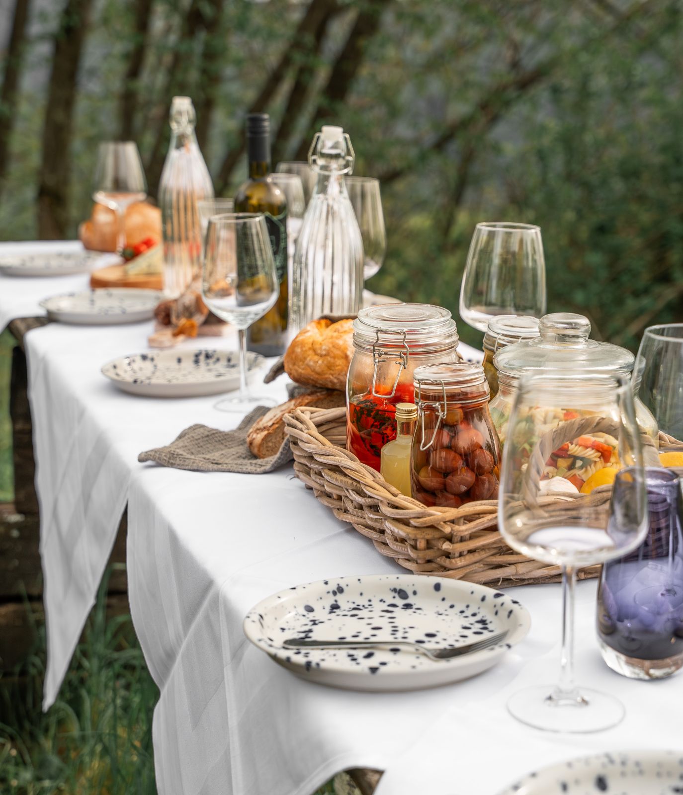 A long outdoor dining table covered with a white tablecloth, set with plates, glassware, bottles, and a basket of jarred foods, surrounded by trees.
