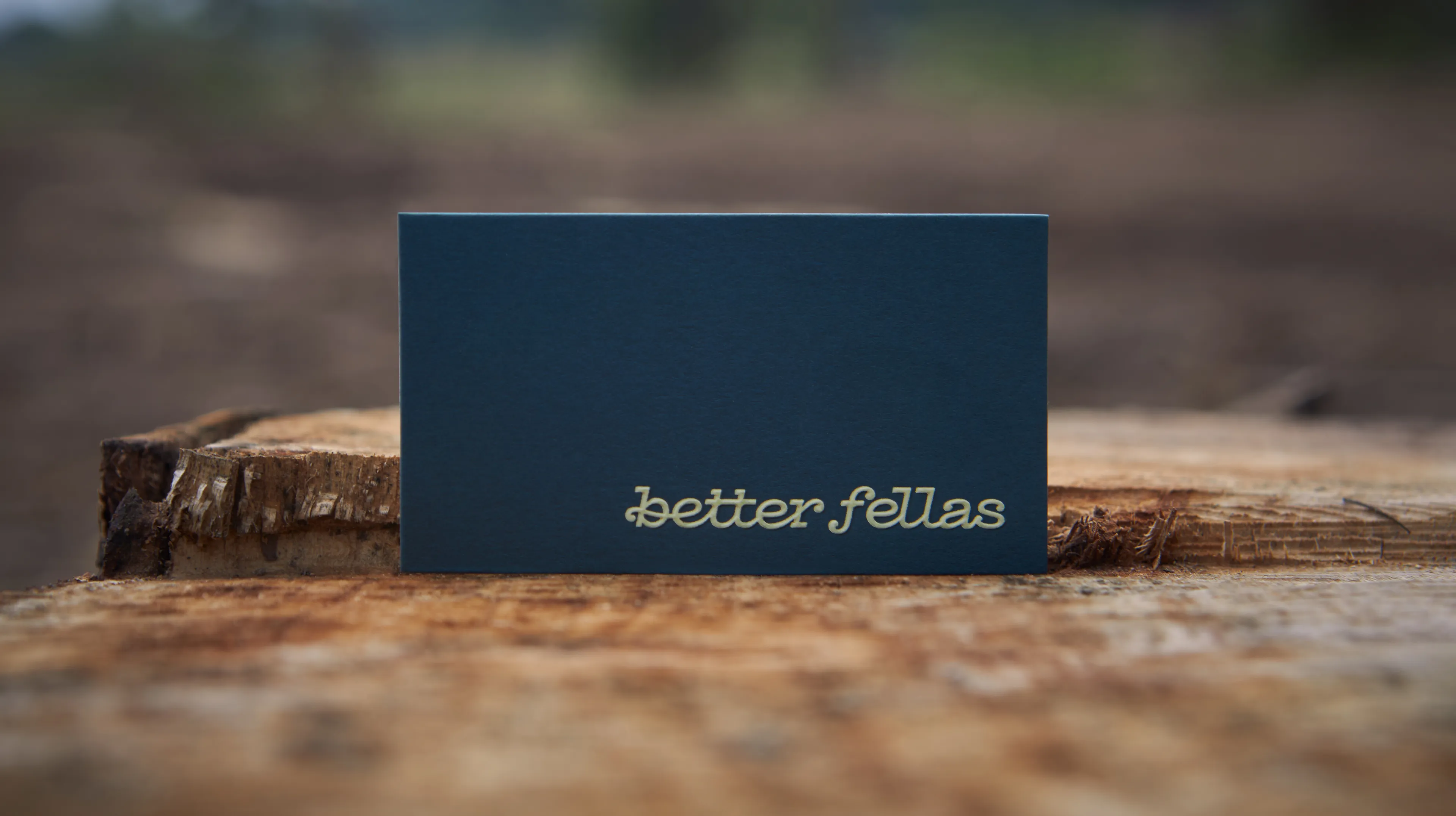 Better Fellas navy blue business card with gold foil script logo propped on a rustic wood stump with blurred outdoor background, designed by Unbound Collective