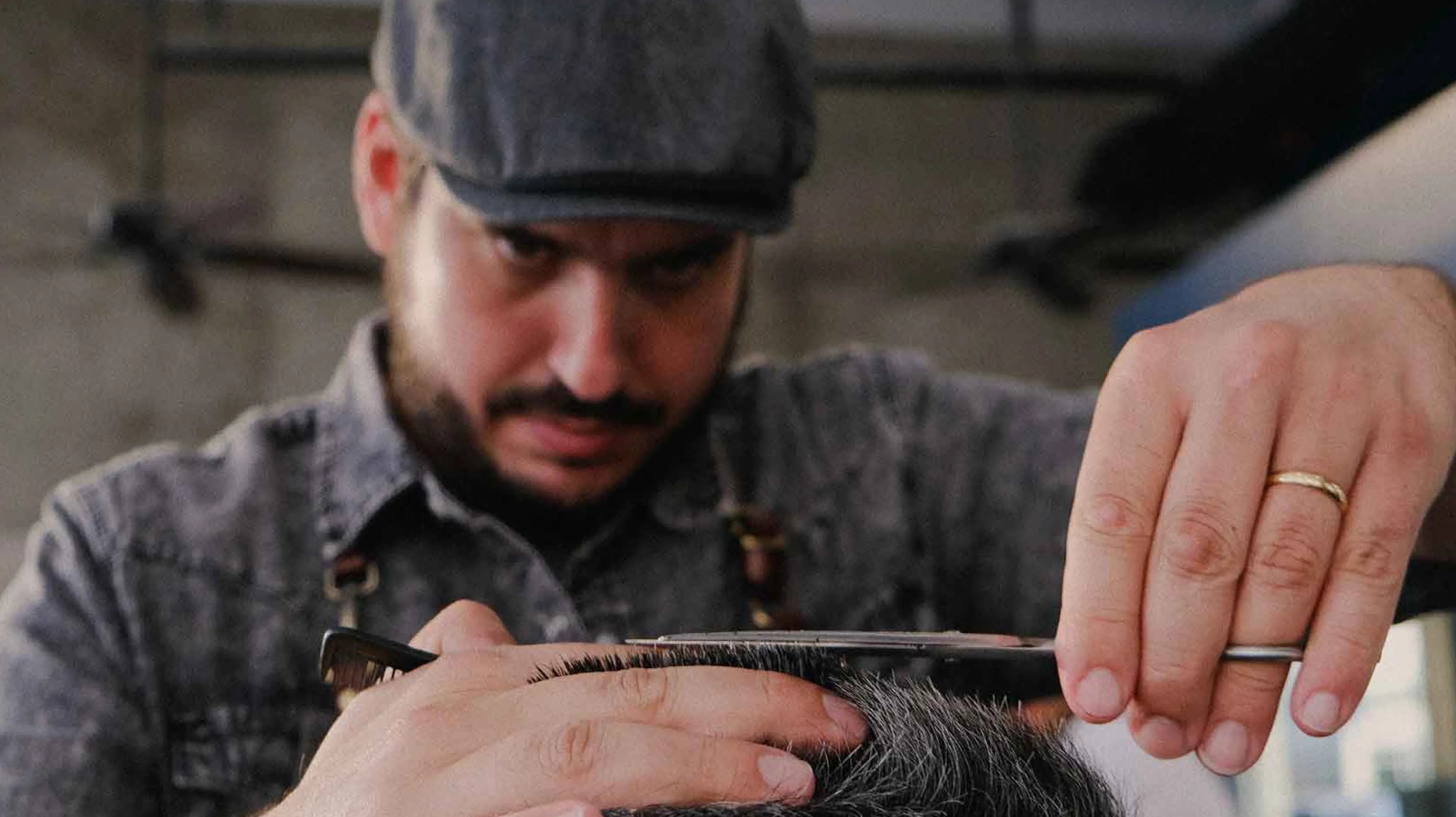 Close-up of a focused barber in a flat cap and denim apron performing a precision scissor-over-comb haircut, brand photography by Unbound Collective for V's Barbershop
