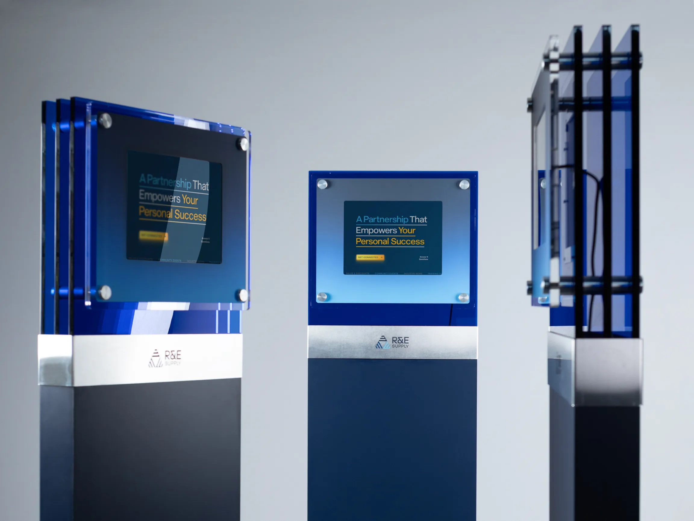 Three R&E Supply branded kiosks in varying sizes with blue acrylic and brushed metal bases, designed by Unbound Collective for trade show and retail environments