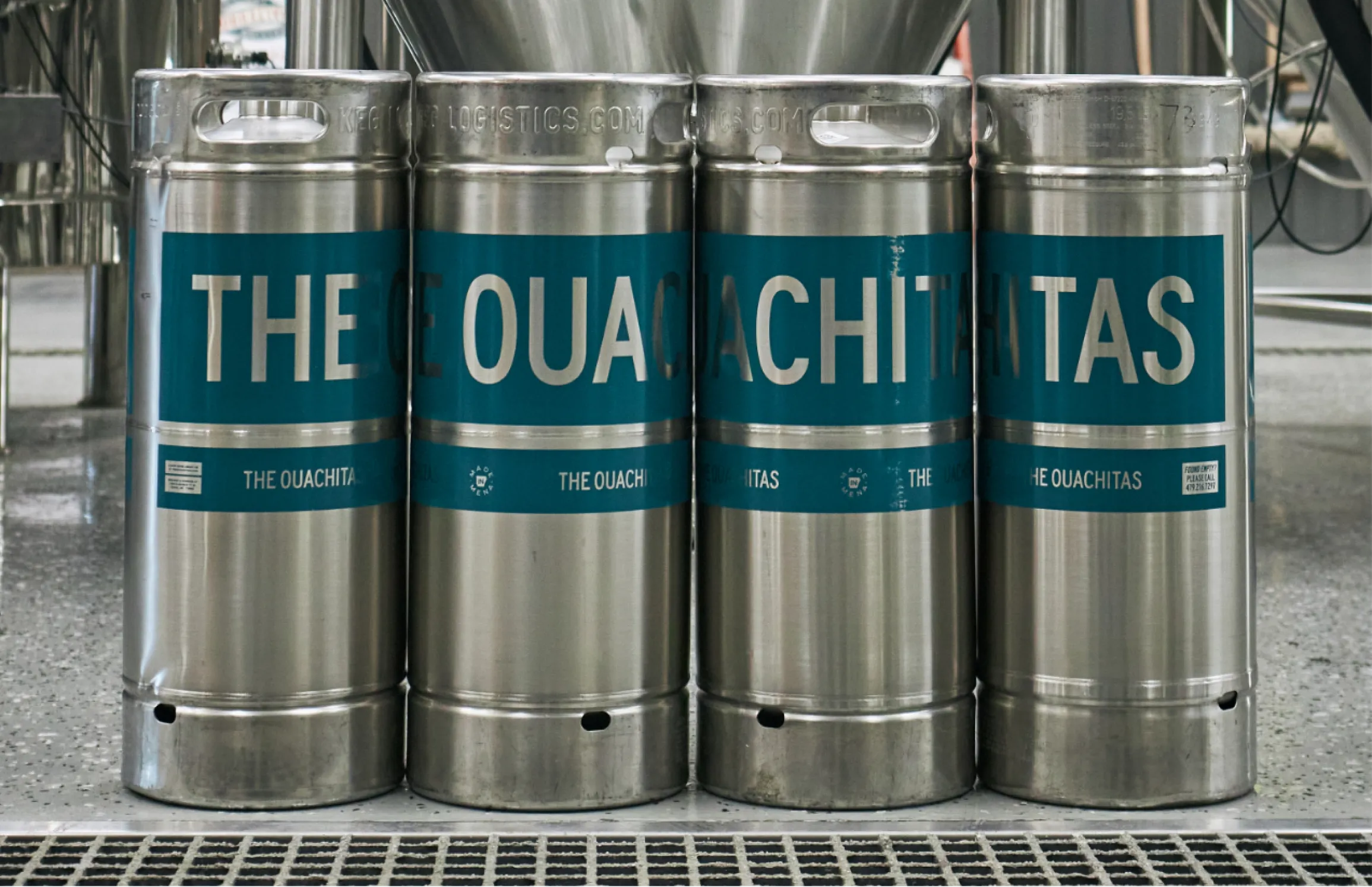 Four stainless steel kegs lined up with teal and white The Ouachitas Brewing branded labels in front of fermentation tanks, photographed by Unbound Collective