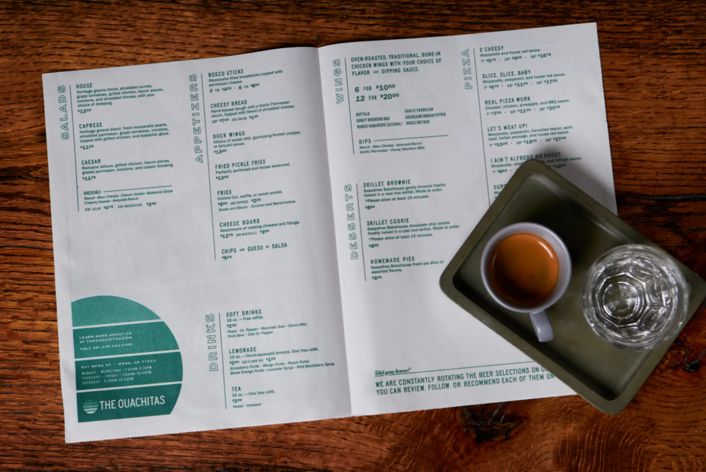 The Ouachitas restaurant menu open spread on a wood table alongside an espresso and water glass showing salads, appetizers, wings, pizza, desserts, and drinks sections in forest green rotated type with teal semicircle contact panel, print and menu design by Unbound Collective