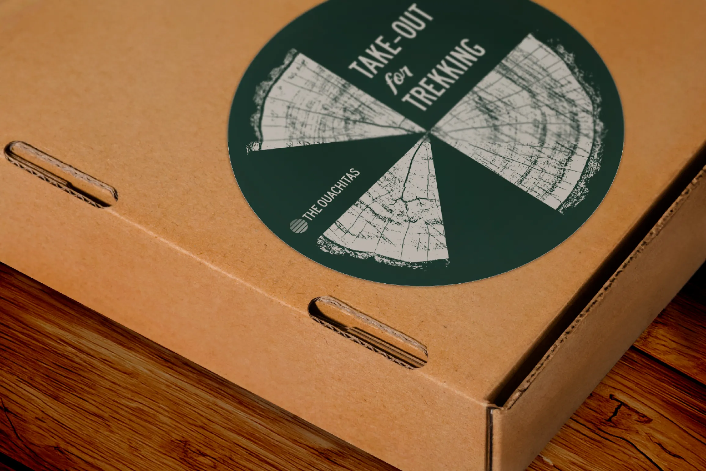 The Ouachitas Take-Out for Trekking pizza box with large circular forest green sticker featuring pizza slice illustrations and The Ouachitas logo on a kraft cardboard box on a wood surface, packaging and graphic design by Unbound Collective