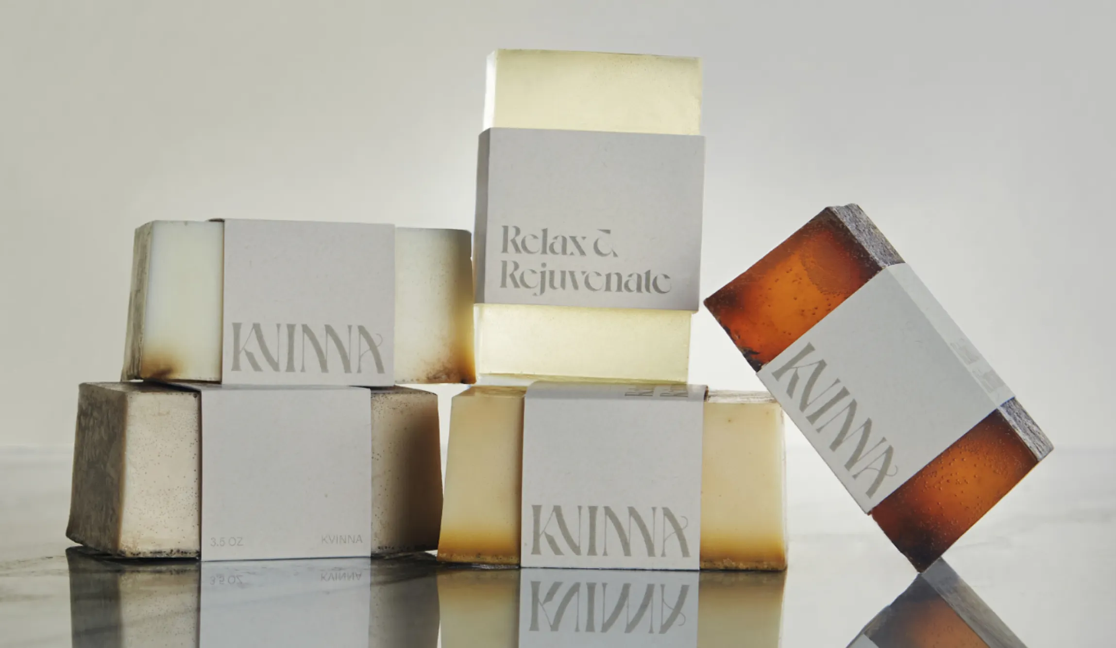Kvinna Studio branded artisan soap bars in gray, amber, and clear varieties with 'Relax and Rejuvenate' paper band packaging and serif logotype on reflective surface, packaging design by Unbound Collective