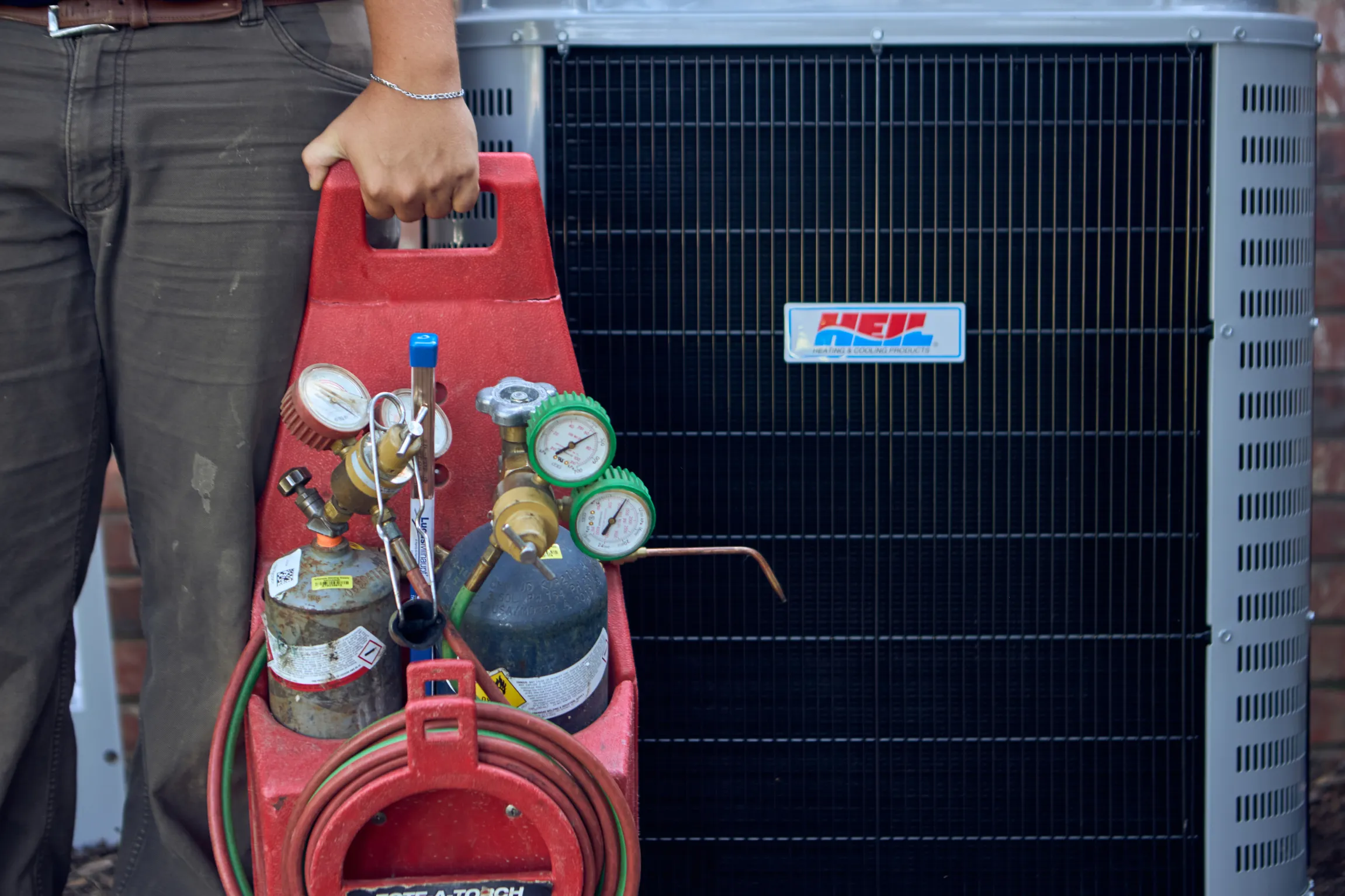 Ski Brothers HVAC technician holding a red refrigerant gauge set beside a Heil outdoor condenser unit at a residential property by Unbound Collective