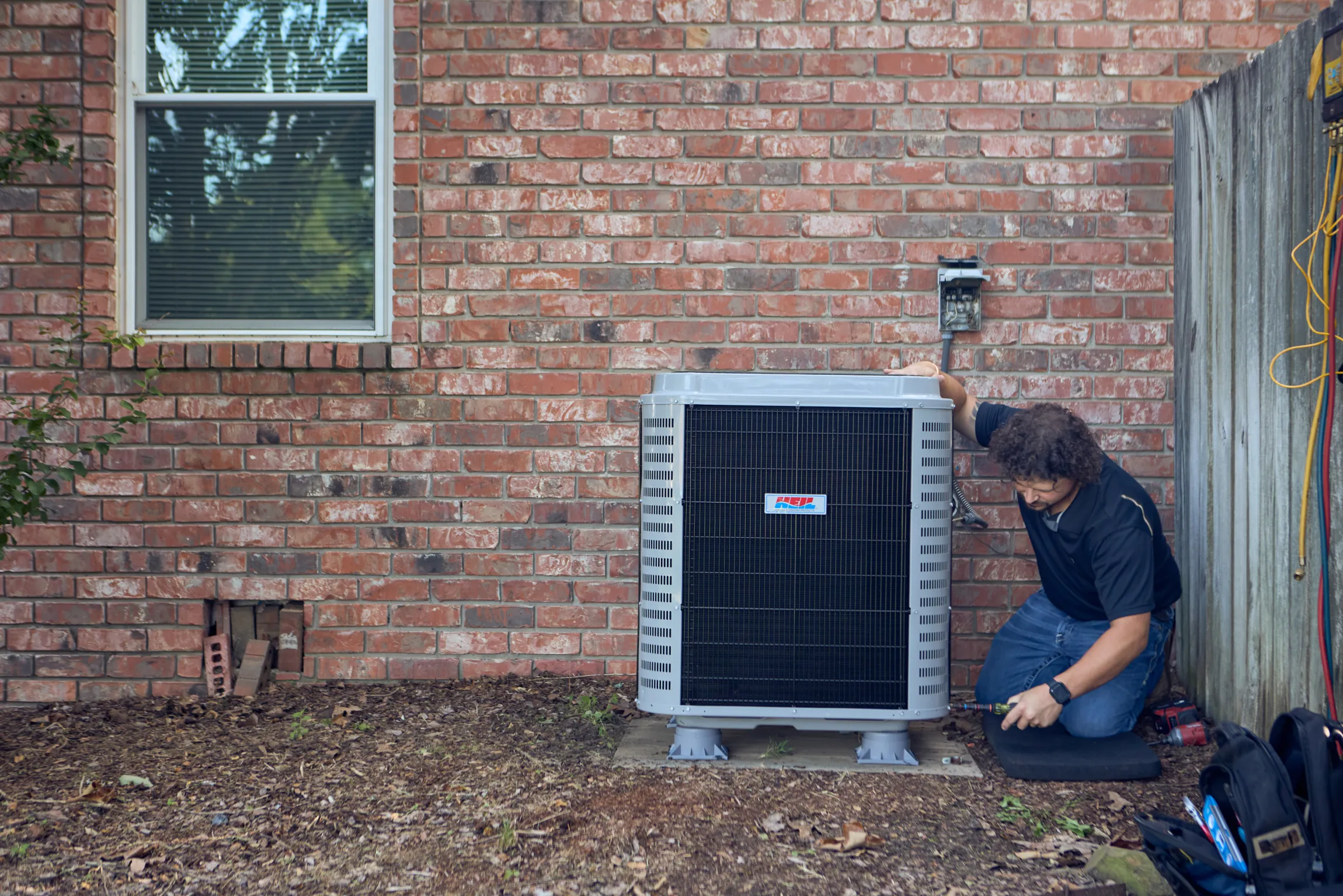 Ski Brothers HVAC technician kneeling beside a newly installed Heil condenser unit against a brick home exterior by Unbound Collective