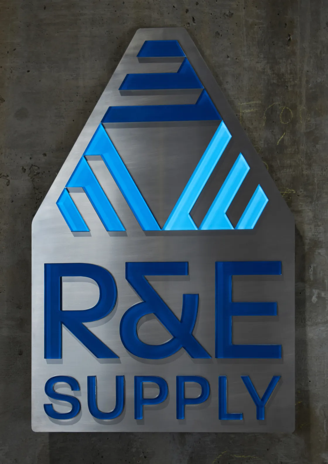 R&E Supply illuminated dimensional logo sign mounted on raw concrete wall featuring brushed stainless steel house-shaped plaque with blue backlit geometric mark and raised blue letter wordmark, signage and brand design by Unbound Collective