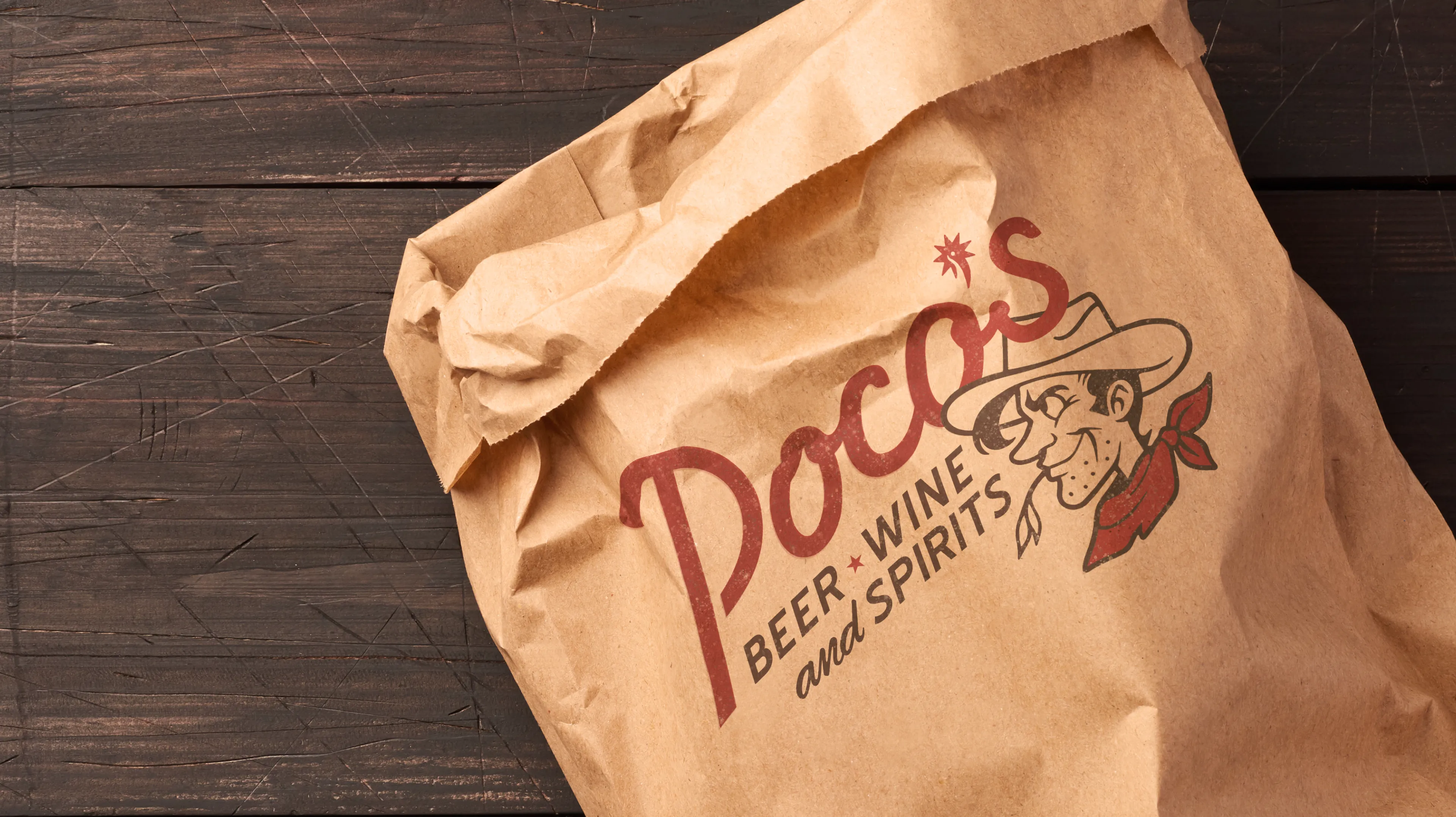 Poco's Beer Wine and Spirits branded kraft paper bag with vintage cowboy logo and script lettering on dark wood surface, packaging designed by Unbound Collective