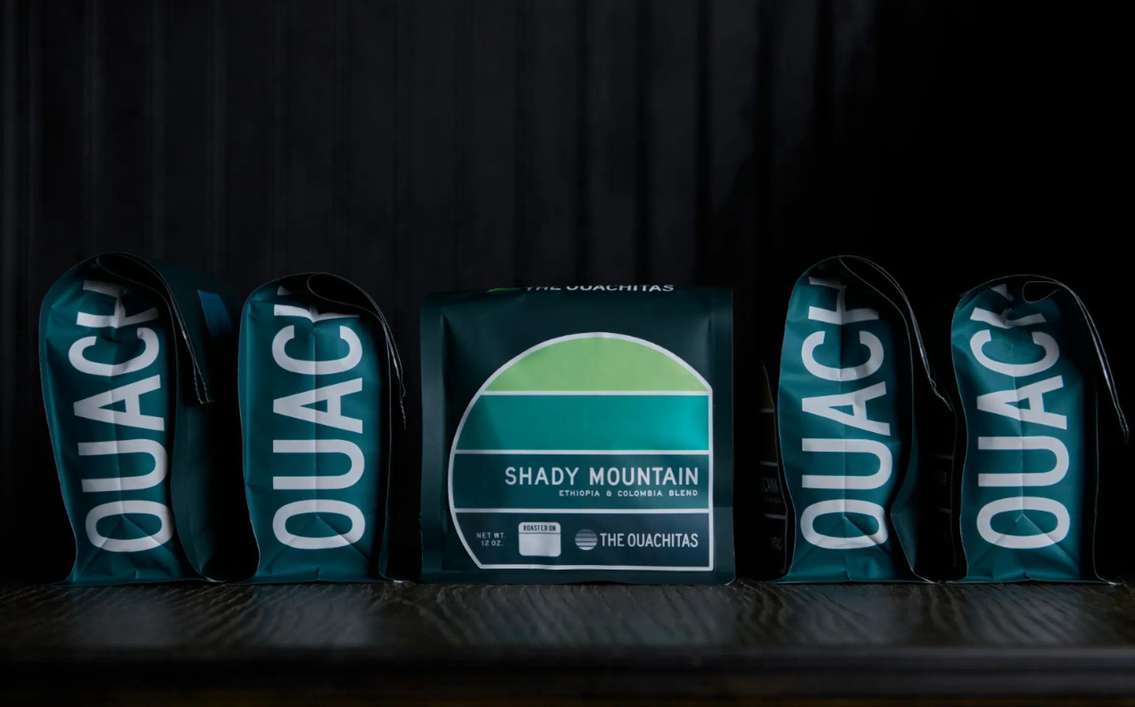 The Ouachitas Shady Mountain Ethiopia and Colombia blend 12oz coffee bags in teal with retro sunrise label displayed in a row on dark background, packaging designed by Unbound Collective