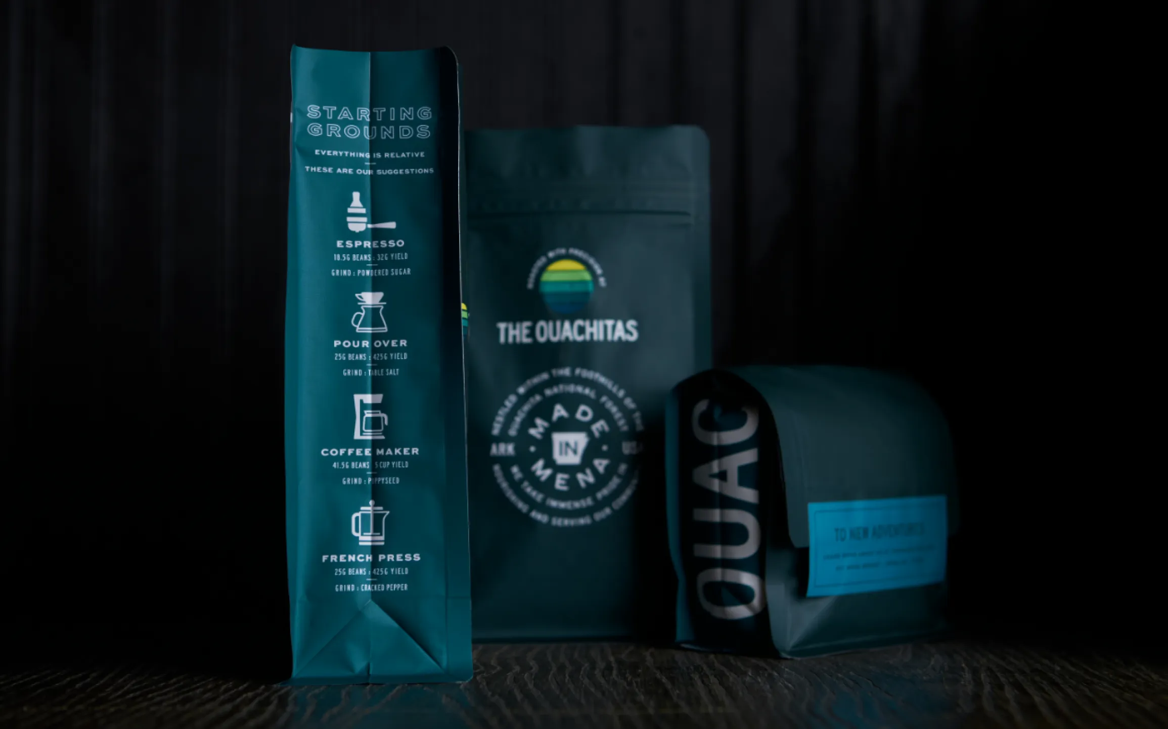 The Ouachitas Starting Grounds teal coffee bag with espresso, pour over, coffee maker, and French press brewing guide alongside Made in Mena and Ouachitas branded bags, designed by Unbound Collective