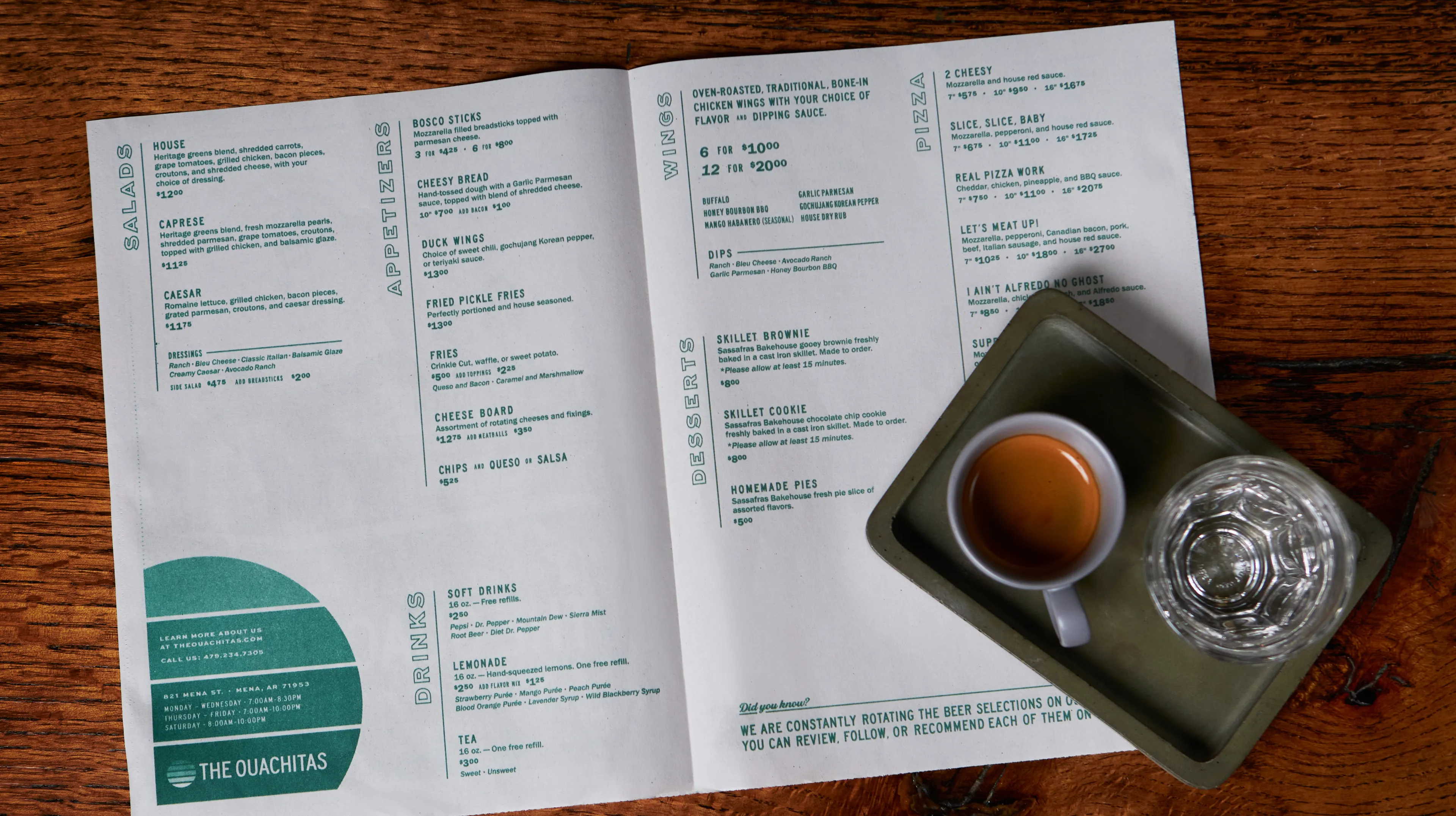 The Ouachitas restaurant menu spread open on a wood table showing salads, appetizers, wings, pizza, desserts, and drinks sections with teal typography, menu design by Unbound Collective