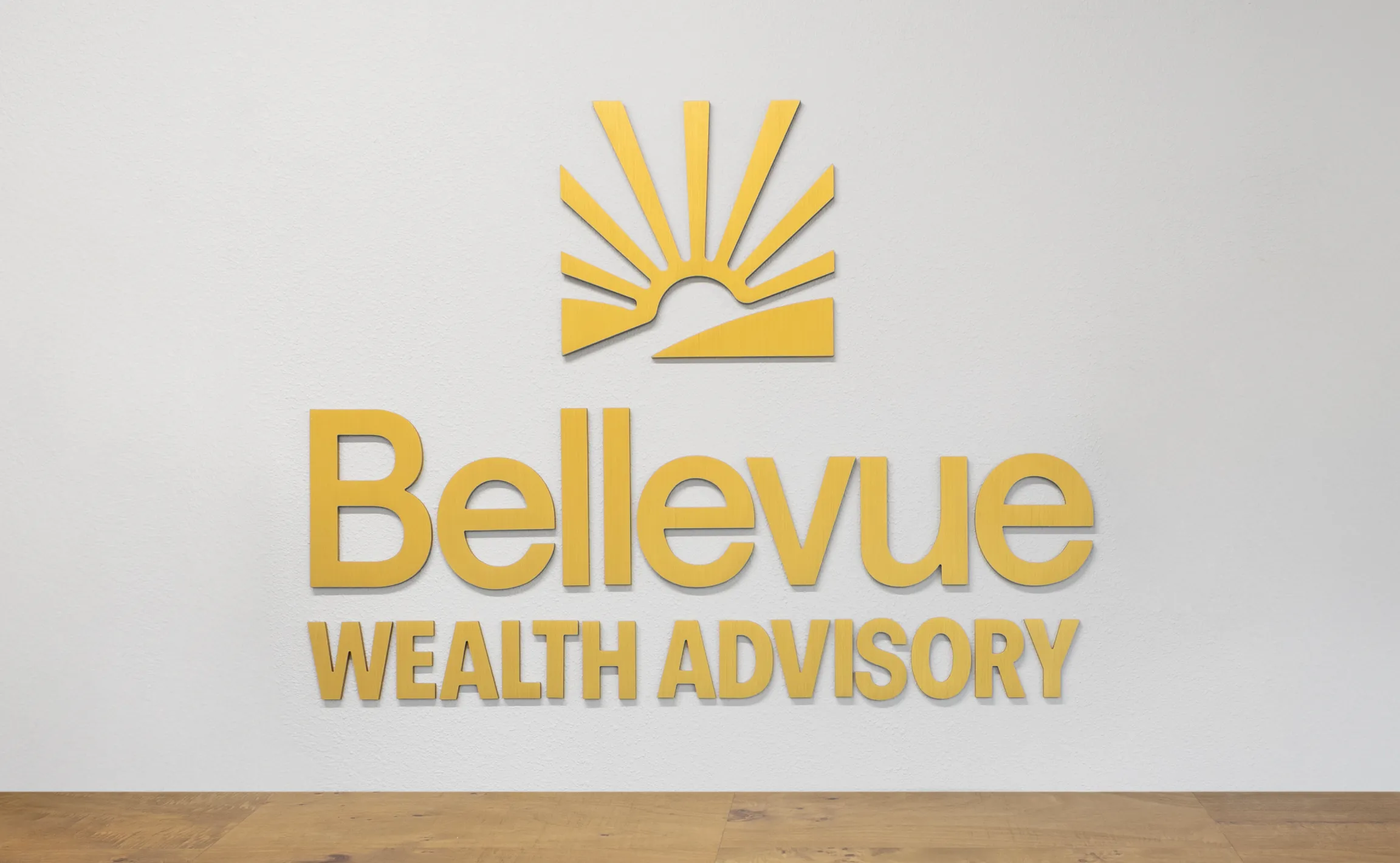 Bellevue Wealth Advisory dimensional lobby sign with gold sunrise logo mark and raised gold letterforms mounted on a white textured wall above hardwood flooring, environmental signage by Unbound Collective