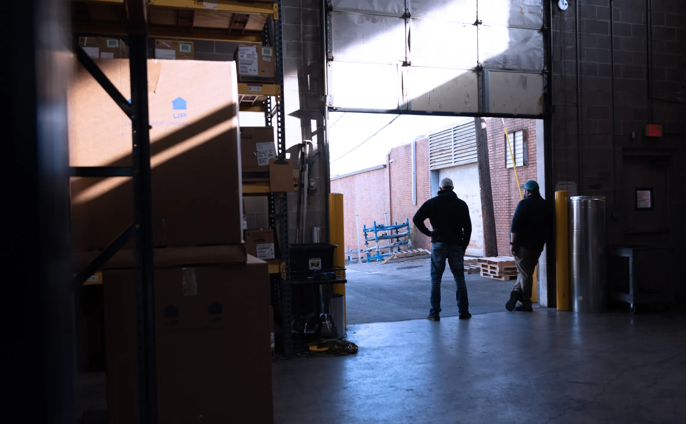 Two R&E Supply warehouse staff standing at an open loading dock bay door with stacked boxes and yellow safety bollards in an HVACR supply warehouse, brand photography by Unbound Collective