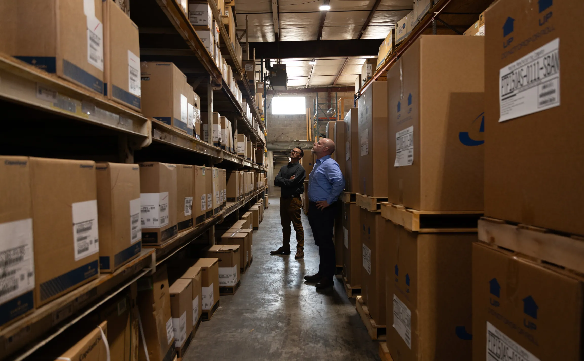 Two R&E Supply team members inspecting HVACR inventory in a warehouse aisle lined floor to ceiling with boxed parts and equipment, brand photography by Unbound Collective