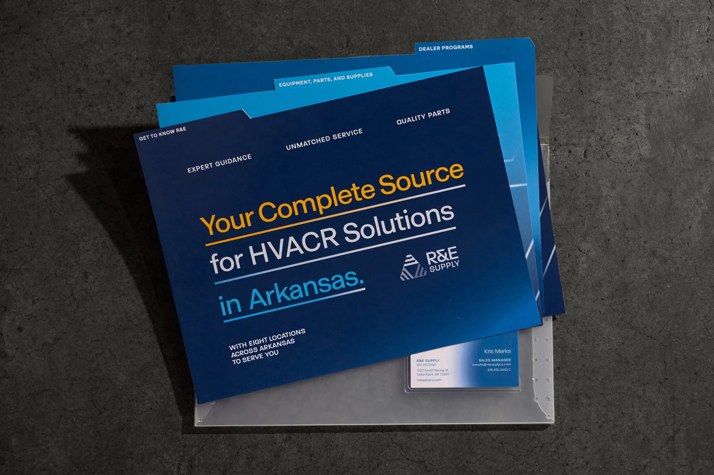R&E Supply full sales collateral flatlay showing navy folder cover with positioning statement, tabbed blue insert sheets for Equipment Parts and Supplies and Dealer Programs, and Kris Marks business card on dark slate surface, print design by Unbound Collective