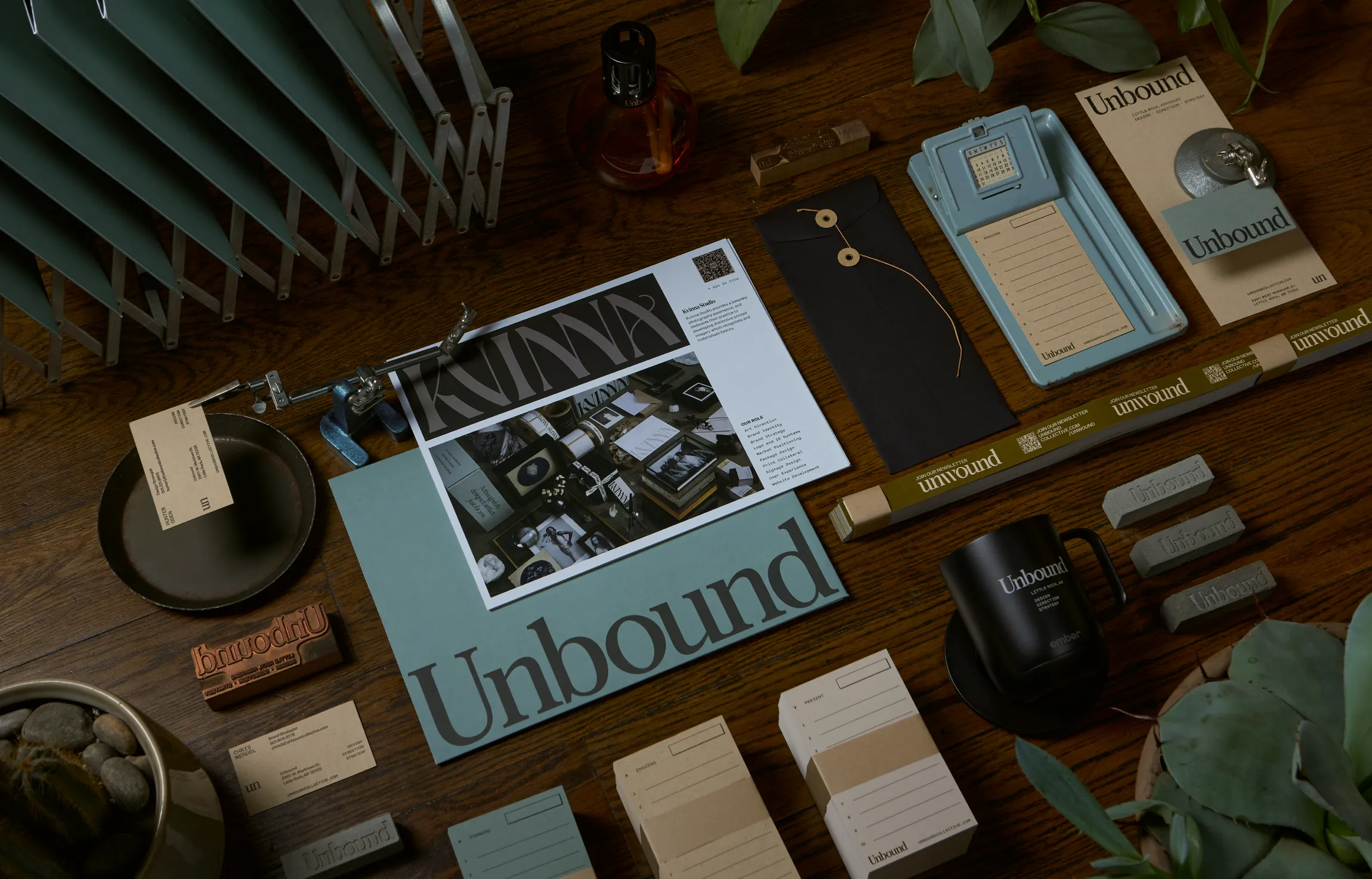 Unbound Collective brand identity collateral flatlay on wood desk featuring stationery, business cards, newsletter prints, branded mug, rubber stamps, and print design materials