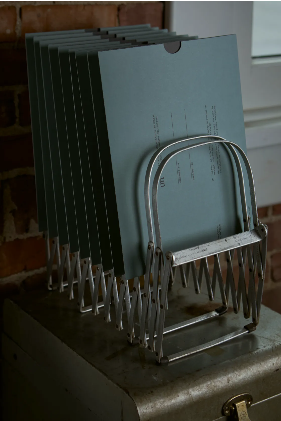 Unbound Collective branded slate blue presentation folders stacked in a vintage metal file rack against a brick wall, print design by Unbound Collective